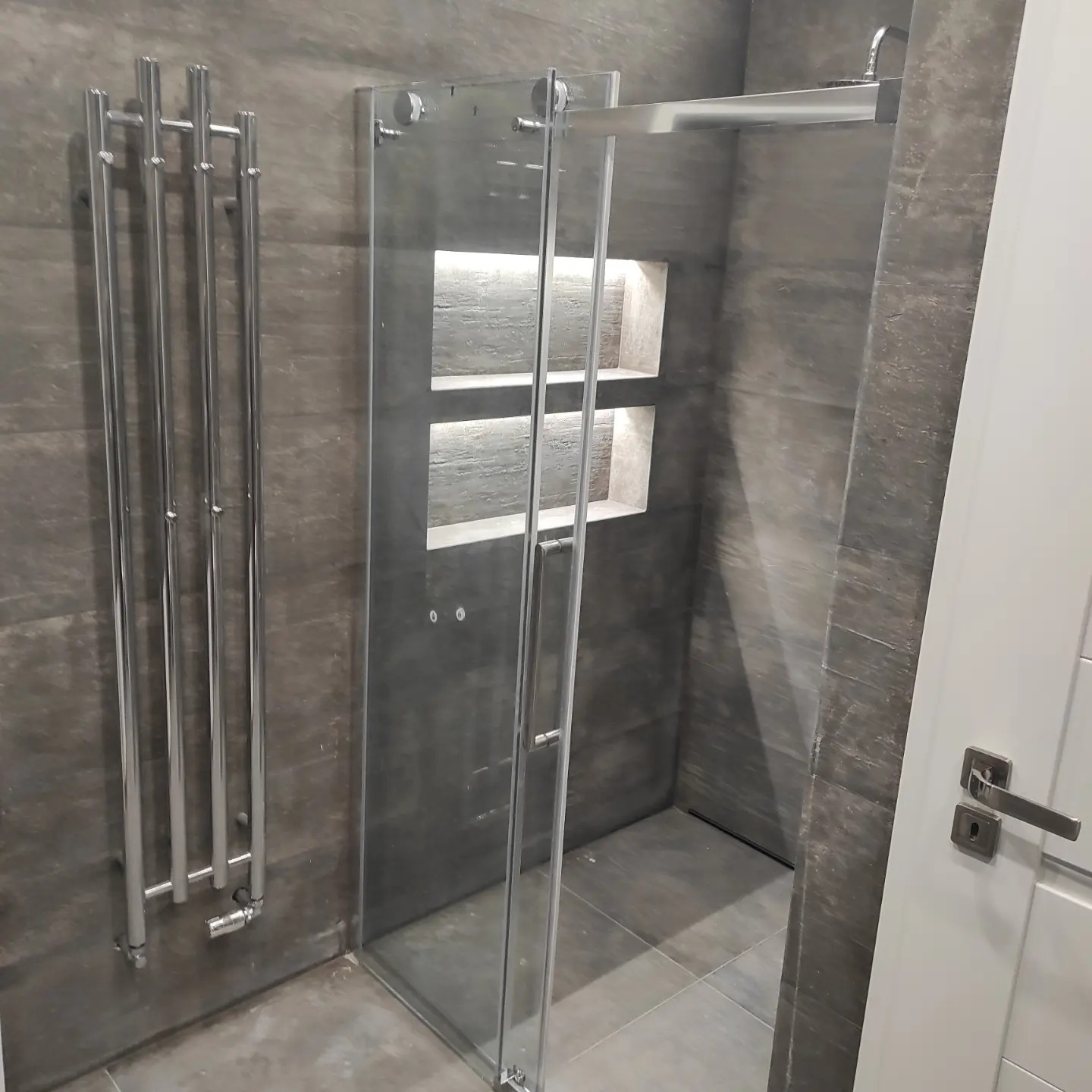 Nowoczesna łazienka z kabiną prysznicową ze szklanymi drzwiami, grzejnikiem drabinkowym i wnękami oświetlonymi w ścianie wyłożonej szarymi płytkami imitującymi kamień.