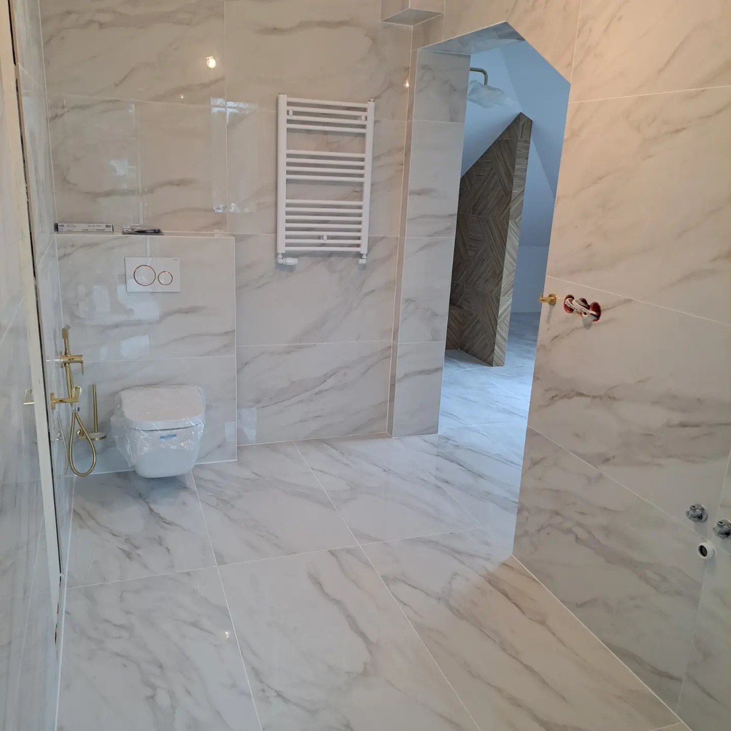 Wykończona łazienka z białymi marmurkowymi płytkami, wisząca toaleta z folią ochronną, grzejnik drabinkowy i nietypowe wejście do kabiny prysznicowej z ukośną ścianą.