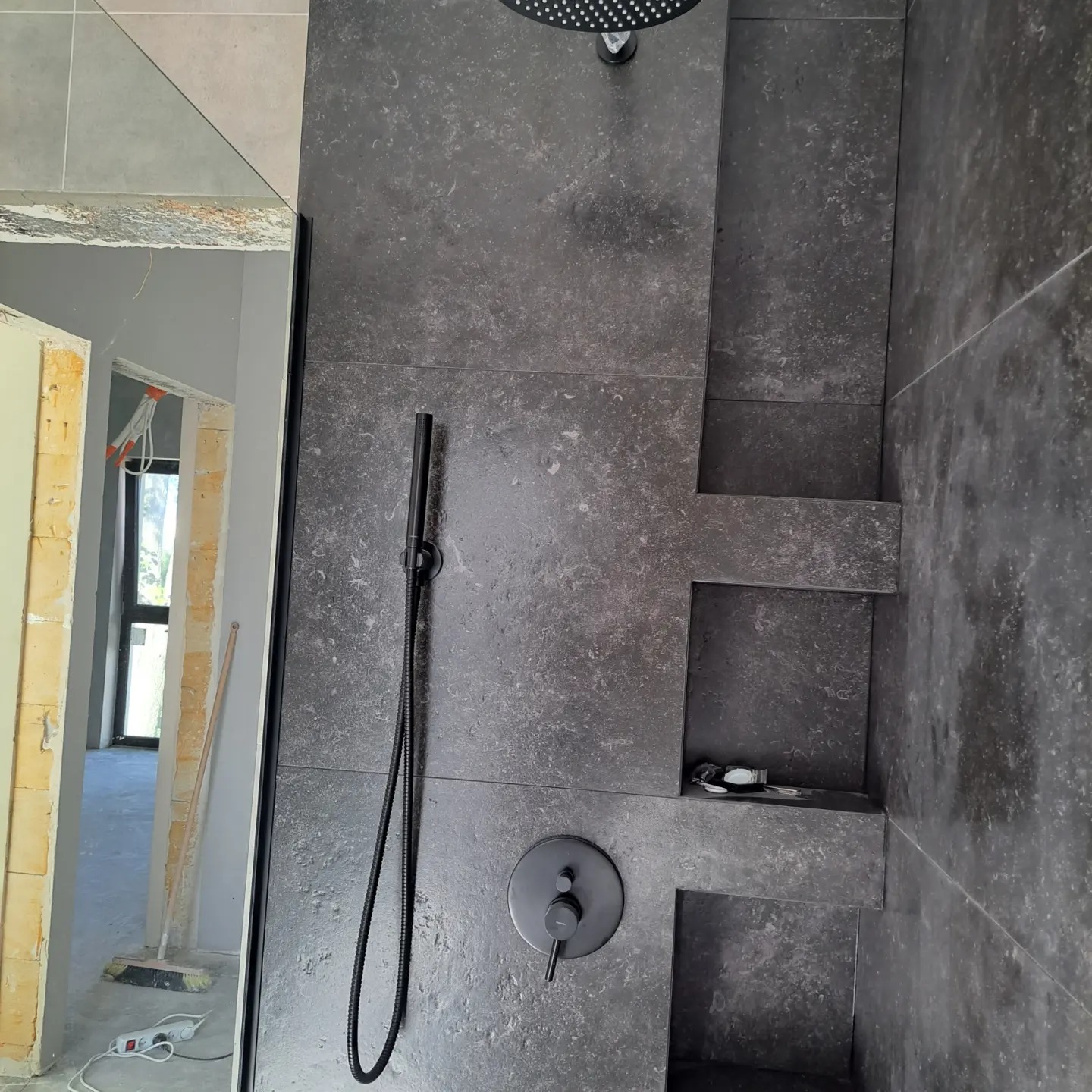Częściowo wykończona kabina prysznicowa z ciemnoszarymi płytkami imitującymi kamień, czarna deszczownica i bateria, wnęki w ścianie na kosmetyki, widoczne niedokończone prace wykończeniowe...
