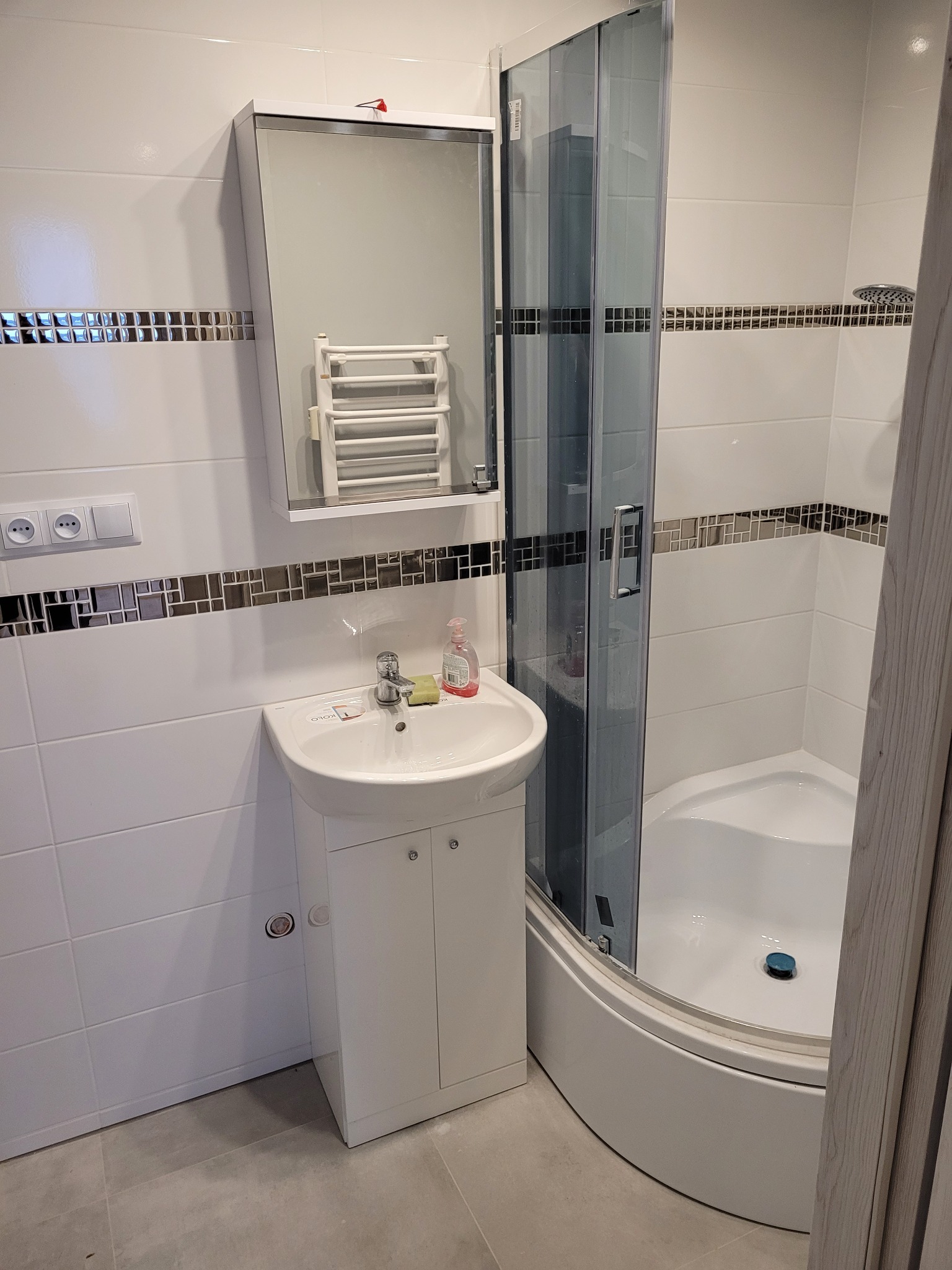 Nowoczesna łazienka po remoncie z kabiną prysznicową z grafitowymi szybami, białą umywalką z szafką, lustrem z grzejnikiem drabinkowym i dekoracyjnym pasem mozaiki na białych płytkach.