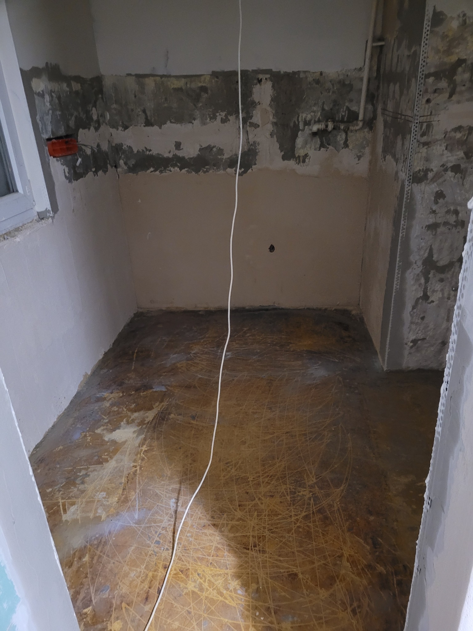 Pomieszczenie w stanie surowym, przygotowane do dalszych prac wykończeniowych, z widocznymi śladami po usuniętej podłodze i częściowo otynkowanymi ścianami. Widoczna puszka elektryczna przy oknie.