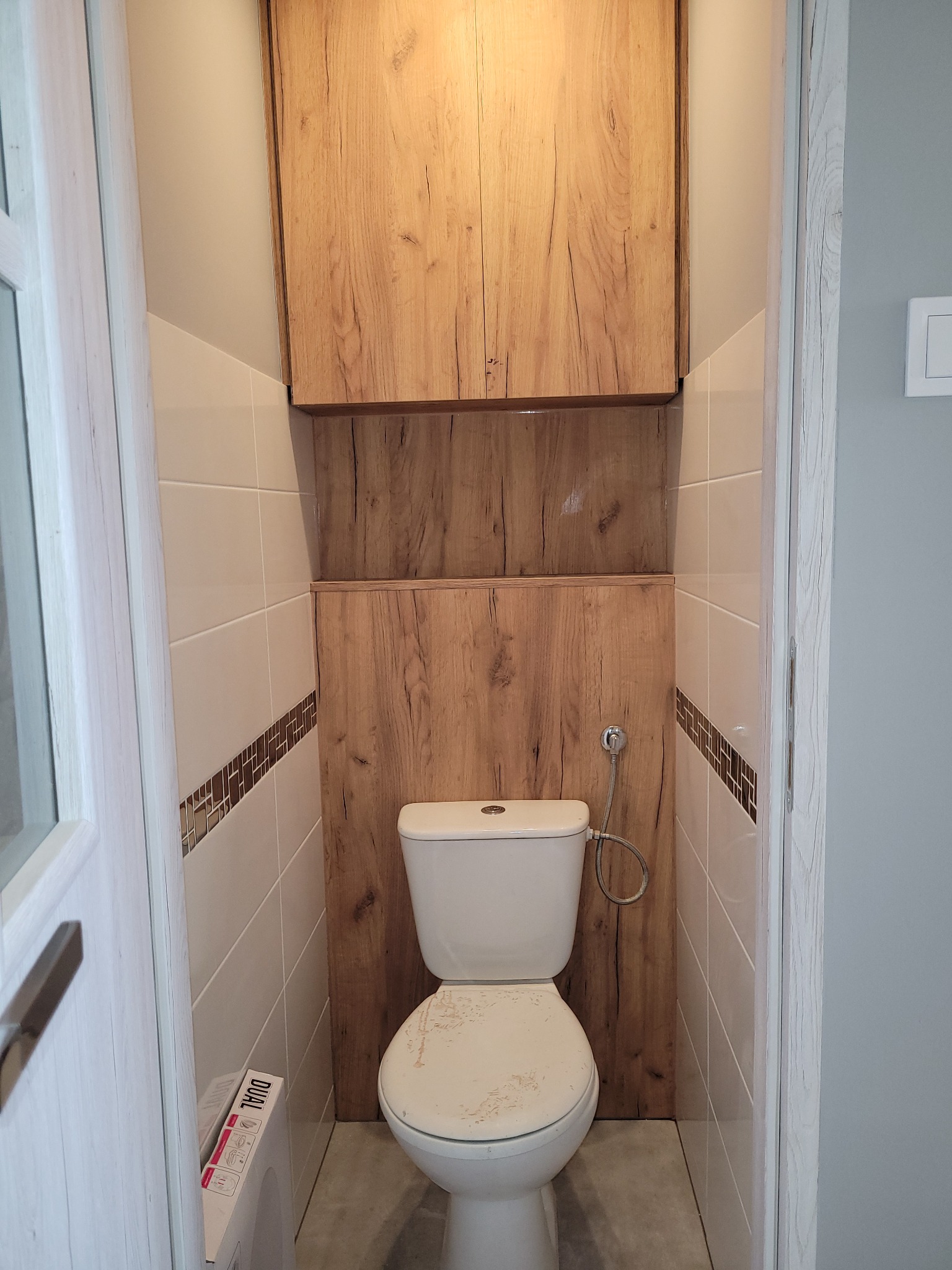 Toaleta z zabudową drewnopodobną, szafka wisząca nad WC, dekoracyjne płytki w pasie na ścianie, widoczne przyłącze wody do spłuczki.