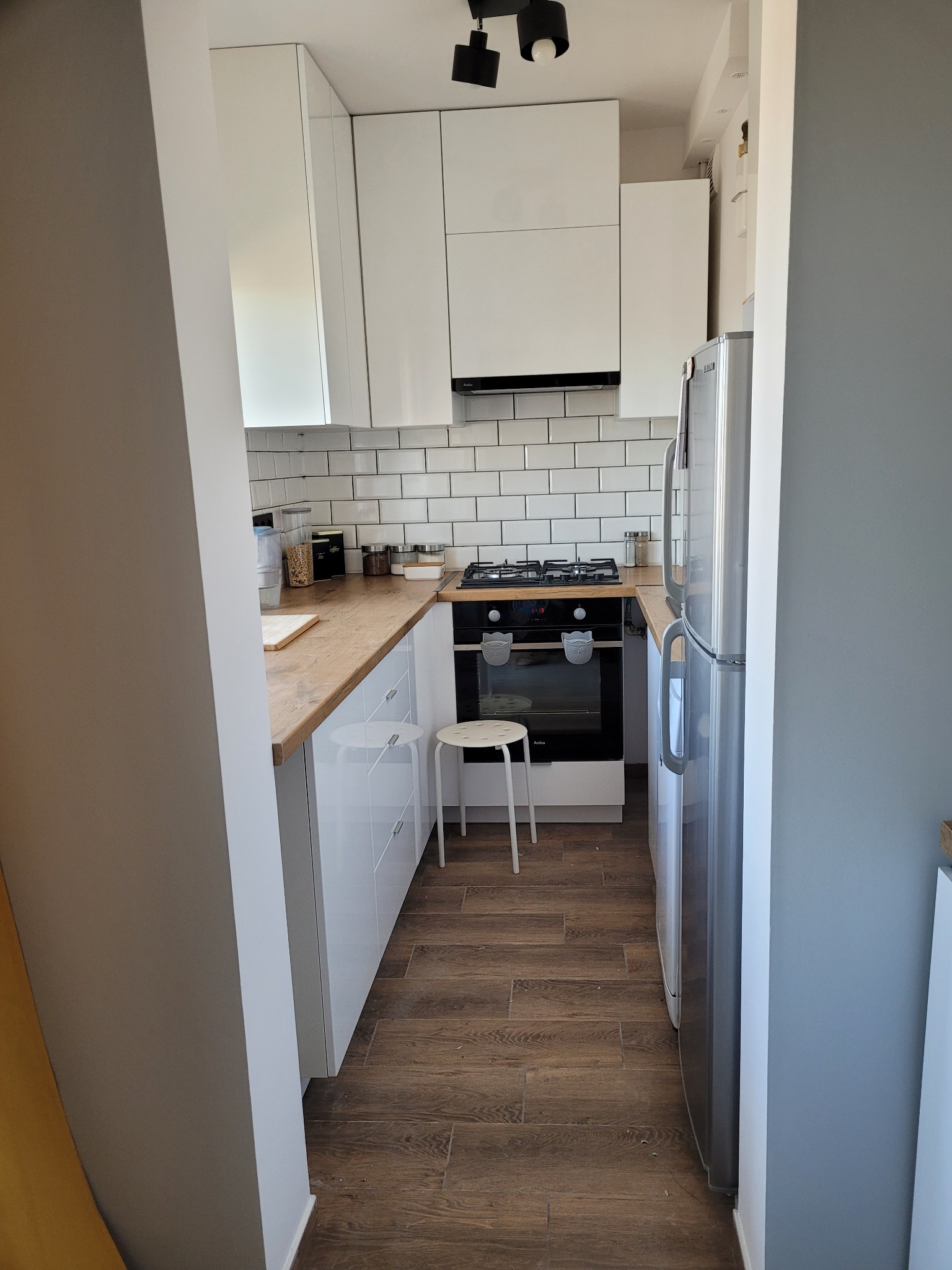 Wąska kuchnia z białymi szafkami, drewnianym blatem i brązową podłogą z paneli, widziana z perspektywy wejścia.