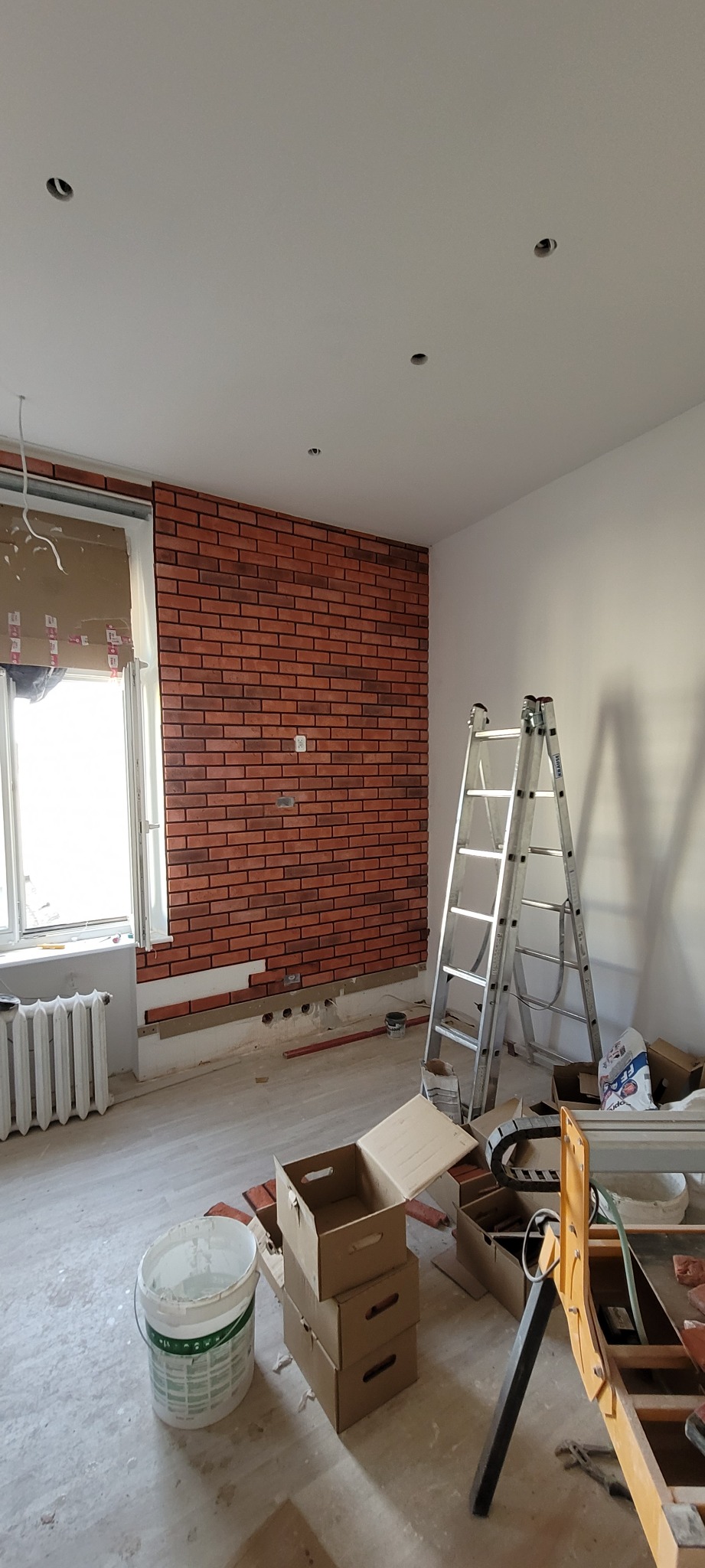 Częściowo wyłożona cegłą ściana w trakcie prac, widoczne puszki elektryczne, drabina, kartony i wiadro z zaprawą.