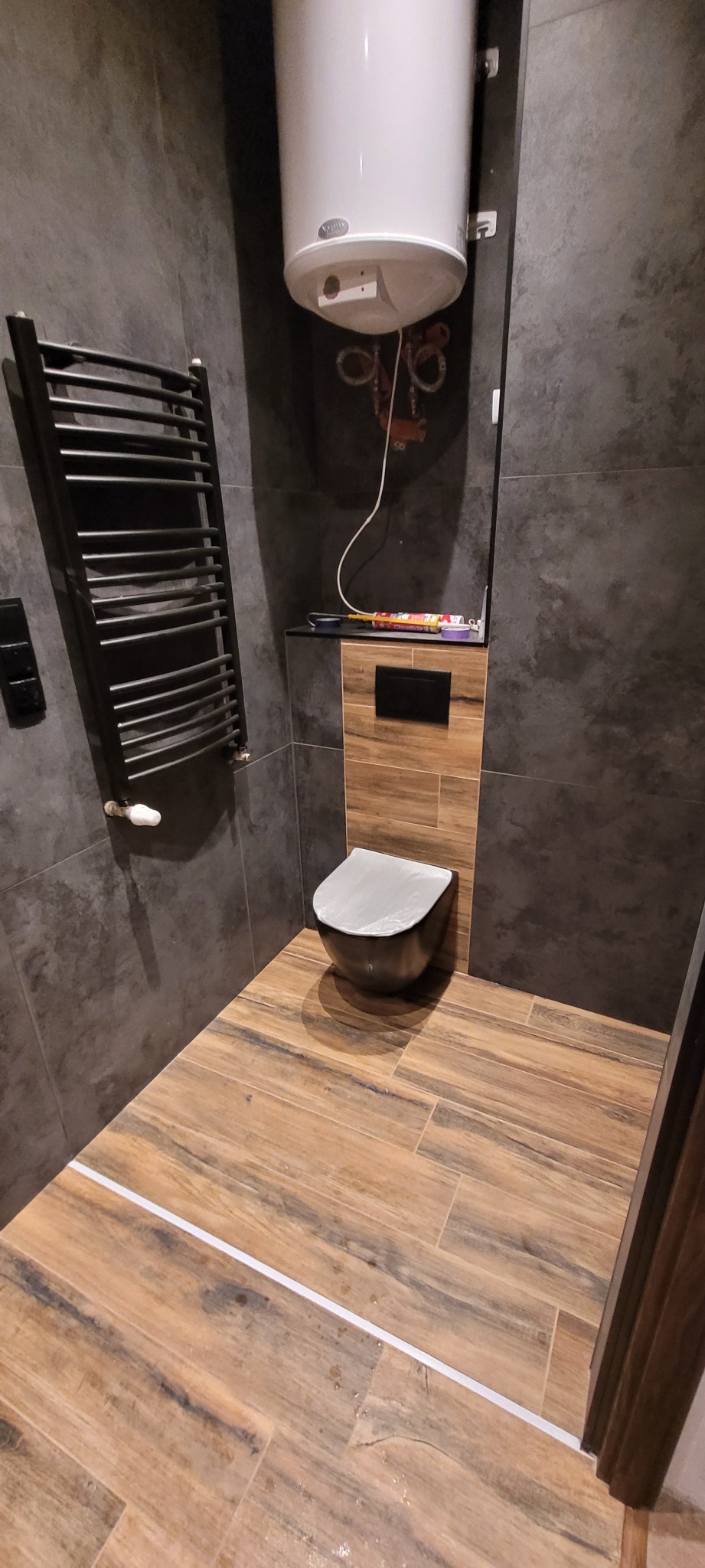 Nowoczesna łazienka z szarymi płytkami imitującymi kamień, drewnopodobną podłogą, wiszącą toaletą z zabezpieczeniem, grzejnikiem drabinkowym i bojlerem. Na półce nad toaletą widoczna tuba z silikonem.