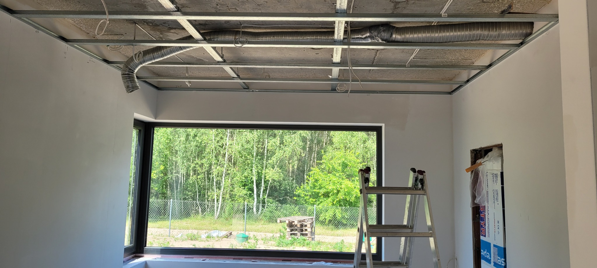 Wnętrze pokoju w trakcie montażu metalowej konstrukcji podwieszanego sufitu, widoczne aluminiowe rury wentylacyjne, drabina oparta o okno z widokiem na zieleń