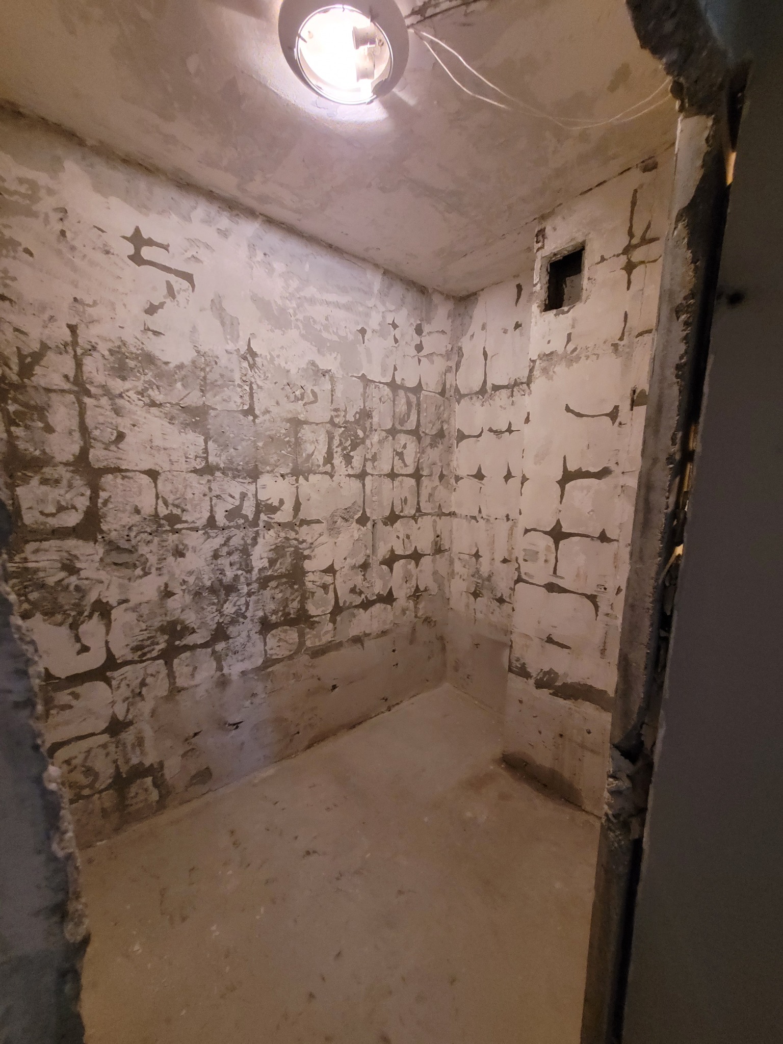 Surowy stan łazienki w trakcie skuwania starych płytek, widoczne nierówności ścian i ślady po kleju, sufit z centralnie umieszczoną lampą.