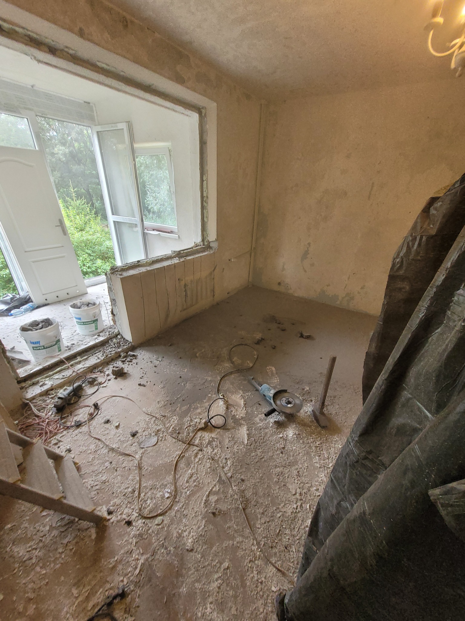 Remont pokoju: wycięty otwór okienny, widoczne narzędzia, gruz i worki z zaprawą, folia ochronna. Przygotowanie do montażu okna.