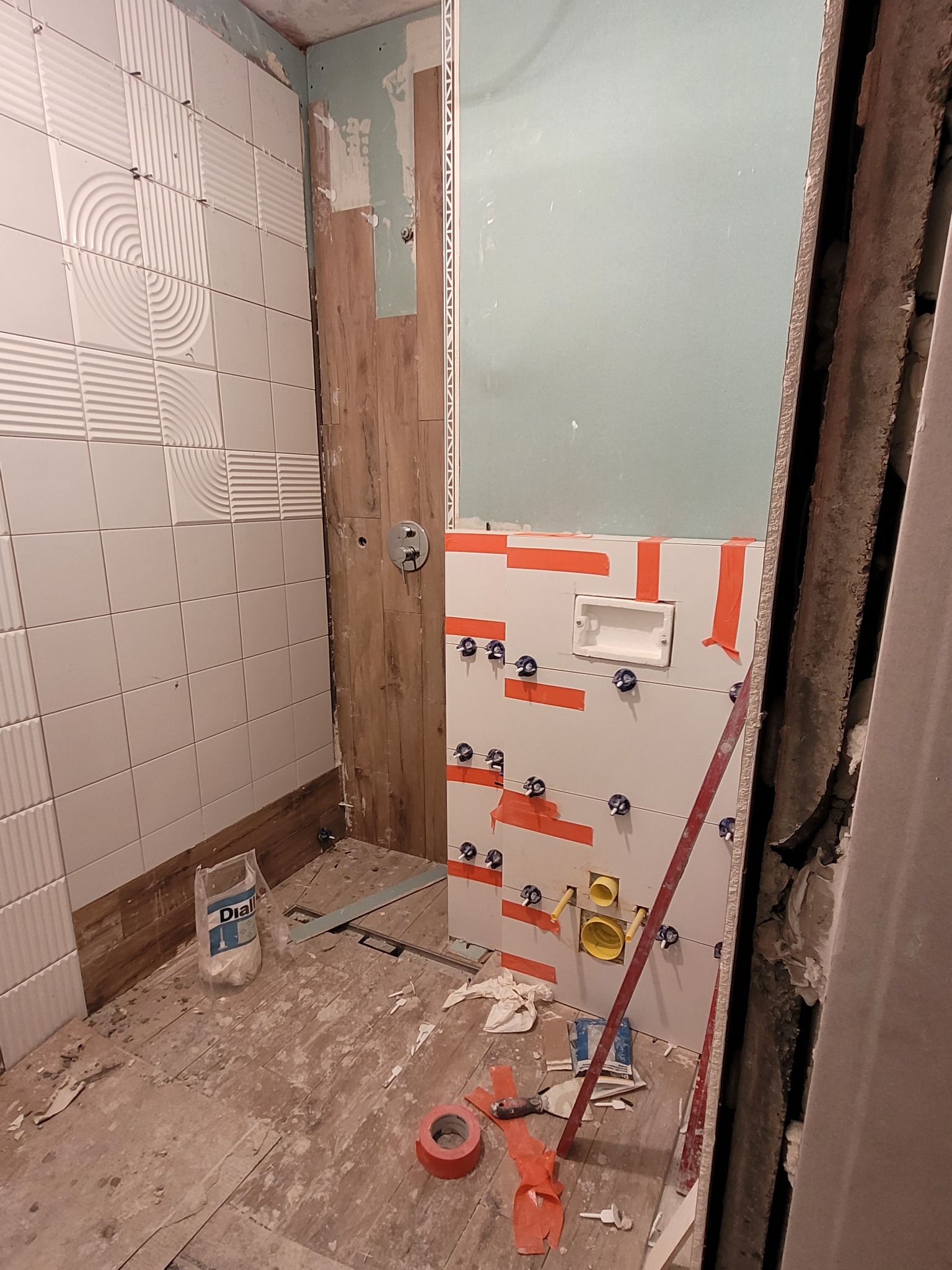 Wnętrze łazienki w trakcie układania płytek ściennych: białe płytki z wzorem 3D na jednej ścianie, płytki imitujące drewno na innej, widoczne poziomice, krzyżyki dystansowe i taśma malarska...