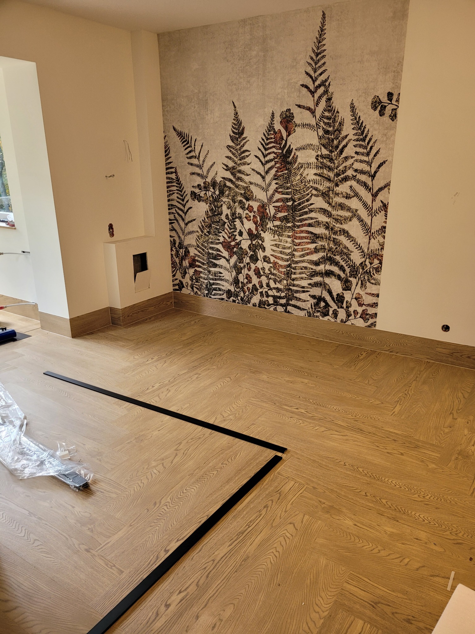 Parkiet ułożony w jodełkę, listwy przypodłogowe w kolorze drewna, ściana z tapetą w motywy roślinne, czarne listwy podłogowe gotowe do montażu.