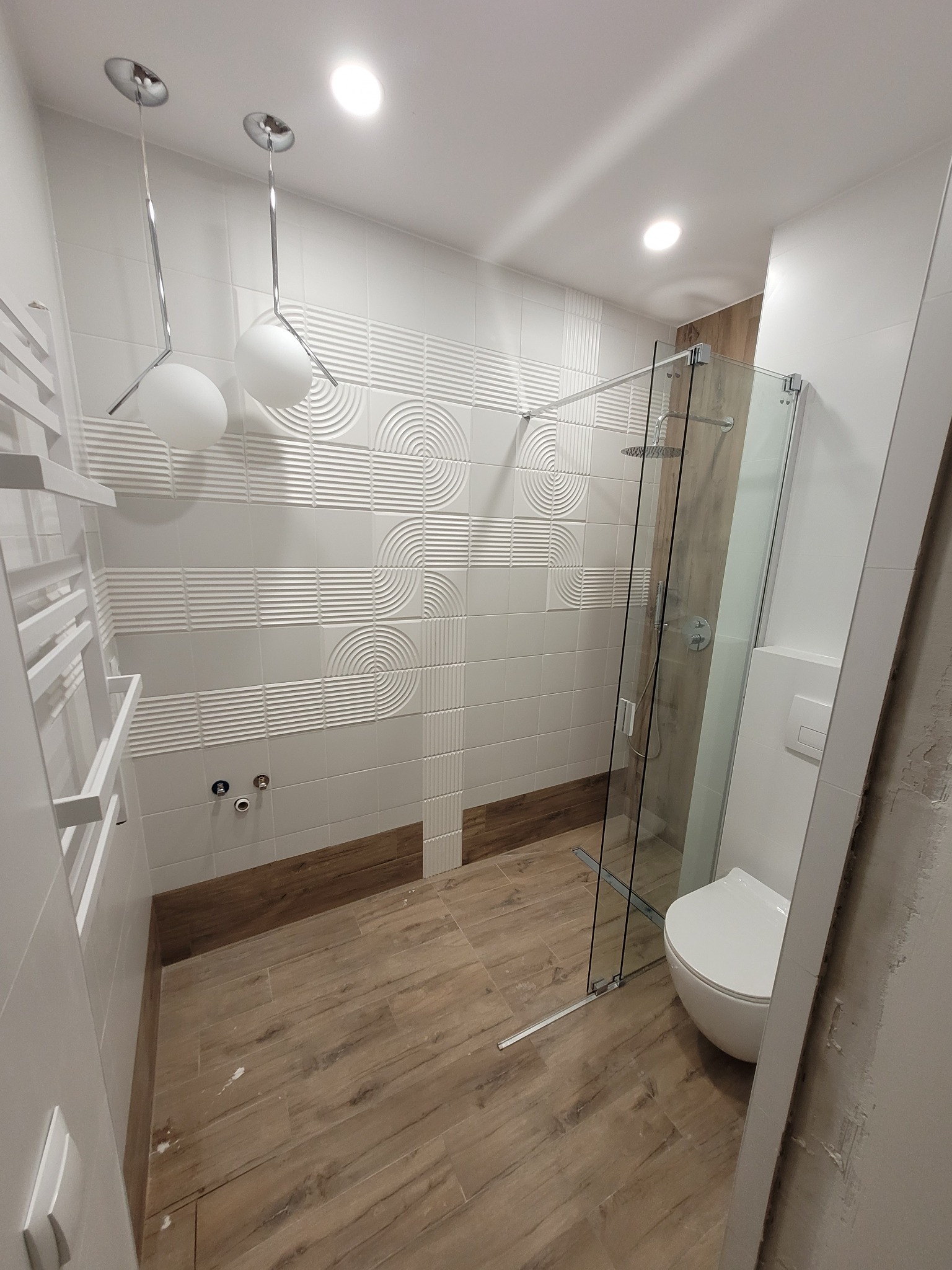 Nowoczesna łazienka z kabiną prysznicową typu walk-in, białymi płytkami z tłoczonym wzorem, drewnopodobną podłogą i wiszącymi lampami kulami.