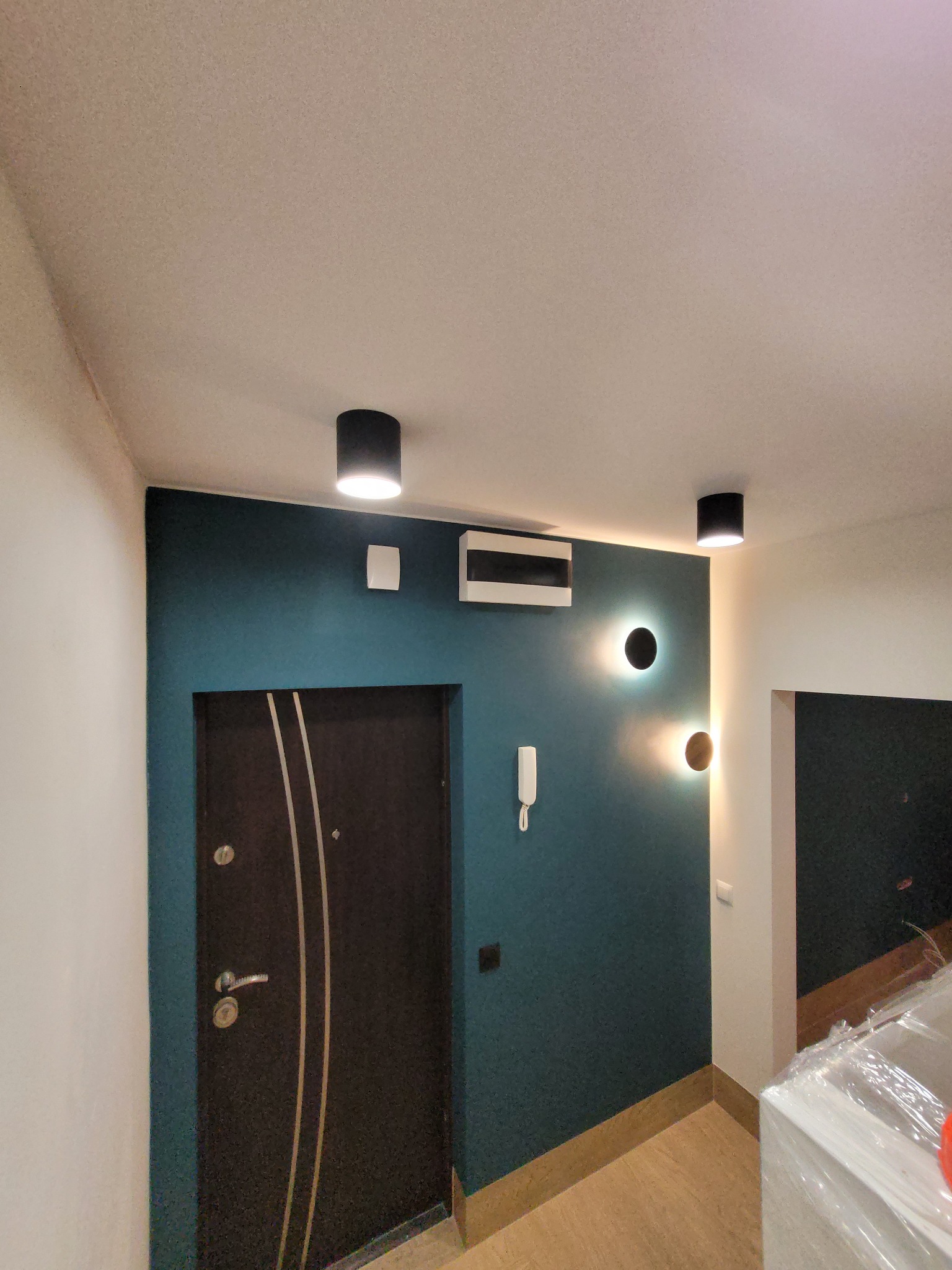 Przedpokój po odświeżeniu: ściana pomalowana na ciemny turkus, czarne lampy sufitowe i ścienne, ciemne drzwi z jasnymi wstawkami.