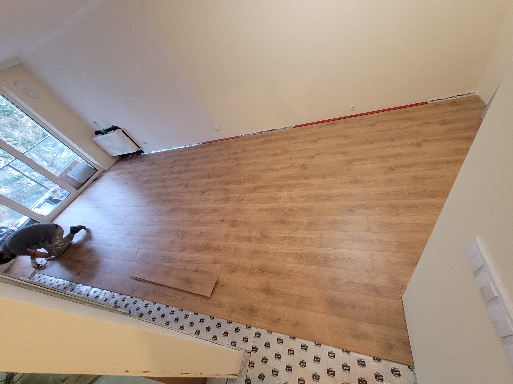 Montaż paneli podłogowych w jasnym pomieszczeniu, widok z góry na pracownika docinającego panele przy ścianie, podkład wyłożony.