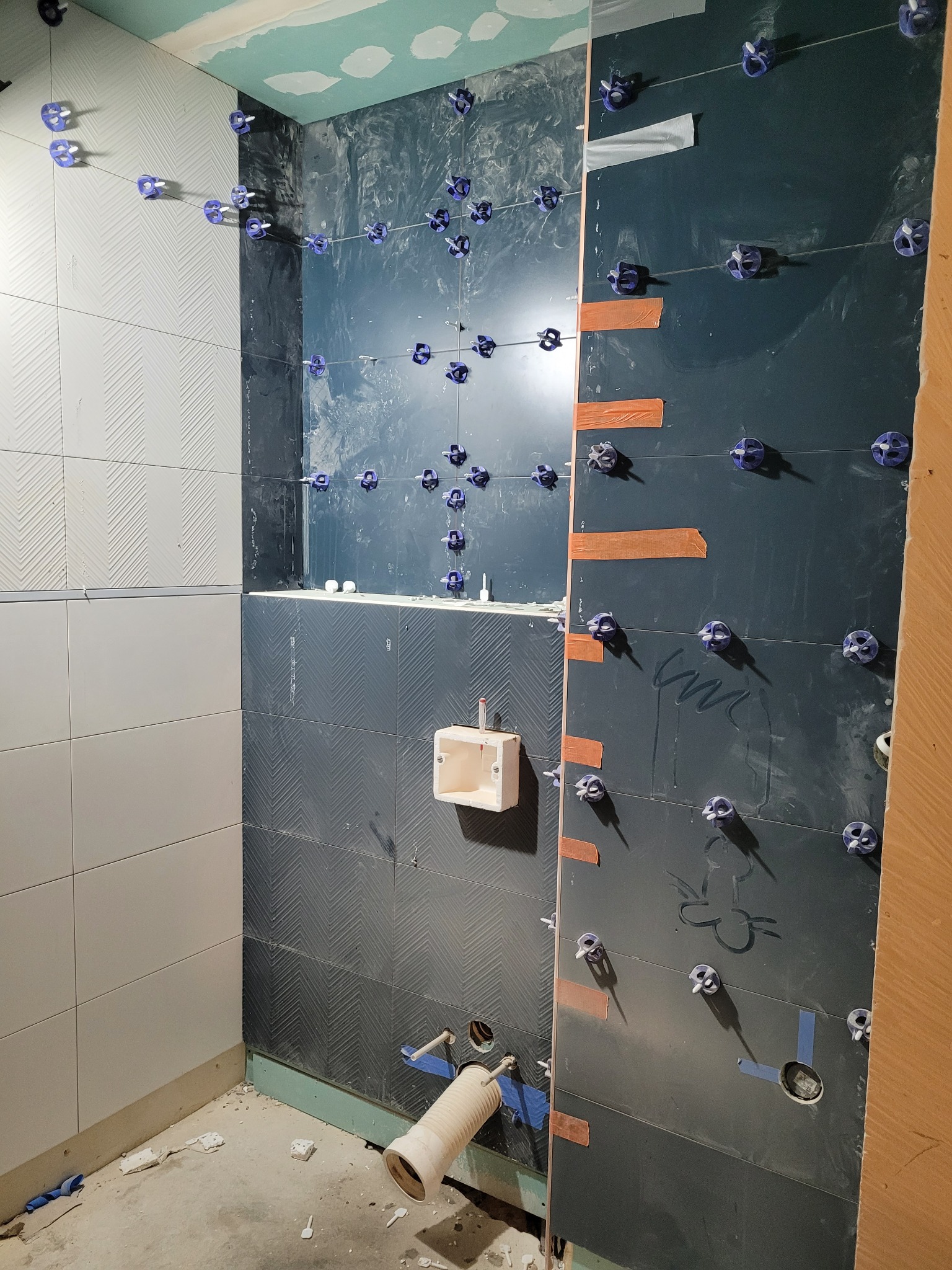 Ściany łazienki w trakcie układania płytek, widoczne krzyżyki dystansowe, różne kolory i wzory płytek, przygotowane otwory na instalacje, oraz prowizoryczne rury odpływowe.