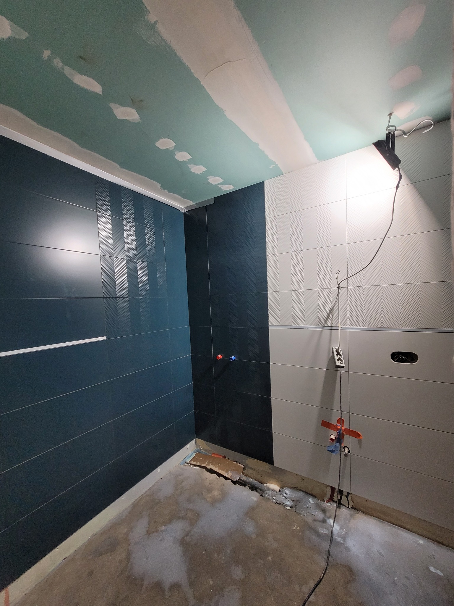 Wnętrze łazienki w trakcie prac: położone płytki w odcieniach granatu i szarości z wzorem chevron, surowy sufit z widocznymi śladami gładzi, podłoga w stanie surowym z resztkami zaprawy, wisząca...