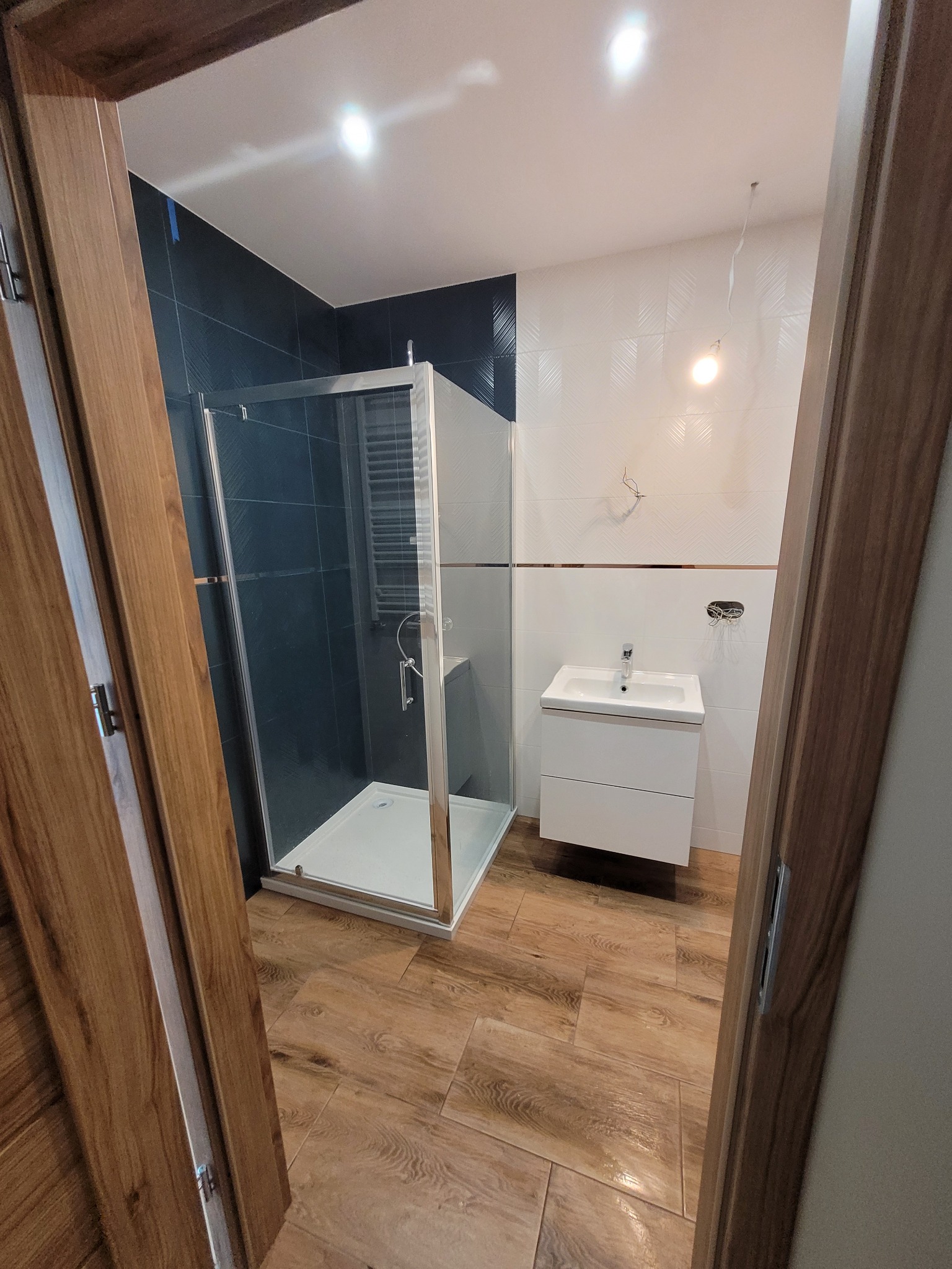 Nowoczesna łazienka z kabiną prysznicową ze szklanymi drzwiami, białą szafką pod umywalką, płytkami w odcieniach bieli i granatu oraz drewnopodobną podłogą, widok zza drewnianej futryny drzwi.