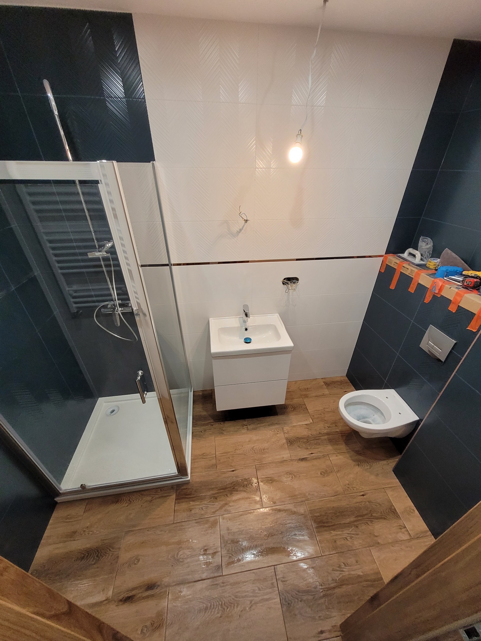 Łazienka w trakcie wykańczania: kabina prysznicowa z chromowaną armaturą, umywalka z szafką, podwieszana toaleta, drewnopodobne płytki na podłodze, biało-granatowe płytki na ścianach z dekoracyjnym...