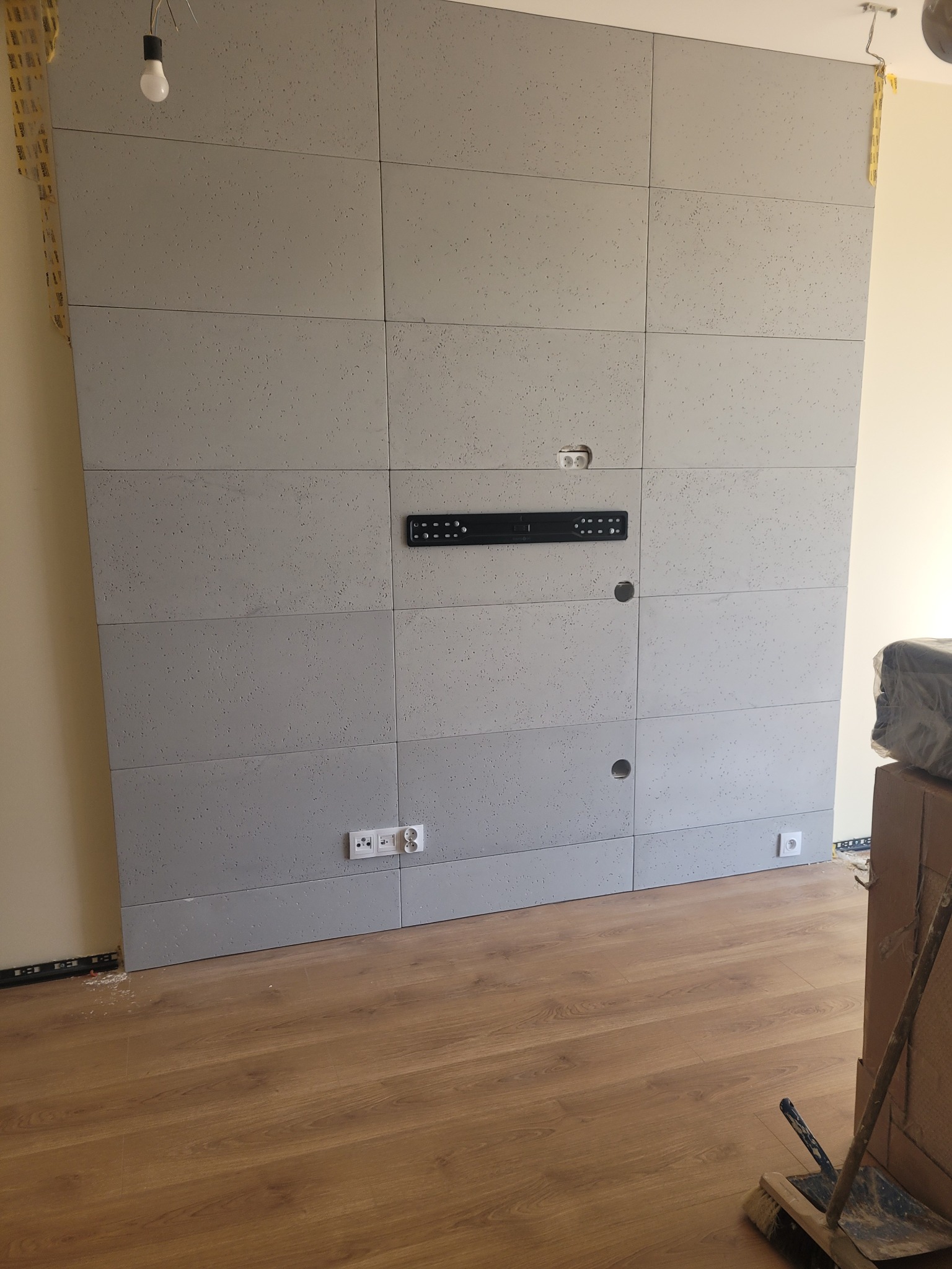 Ściana wykończona szarymi panelami akustycznymi z widocznymi gniazdkami elektrycznymi, uchwytem na telewizor i drewnianą podłogą w trakcie remontu.