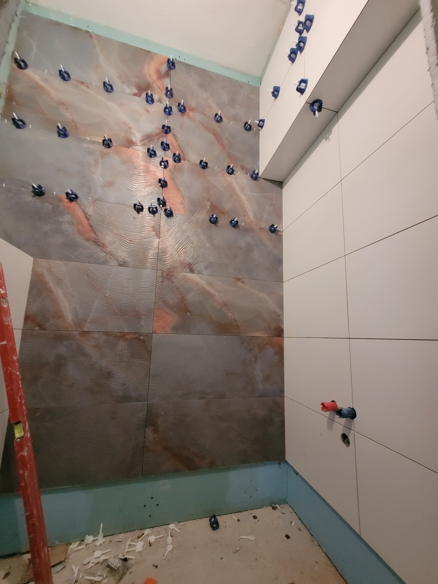 Ściana łazienki w trakcie układania płytek, z widocznymi krzyżykami dystansowymi i poziomnicą, płytki imitujące marmur kontrastują z białymi płytkami, widoczne przyłącza wodne.