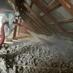 Pracownik w kombinezonie ochronnym aplikuje szarą izolację z dyszy węża na drewnianą konstrukcję poddasza, widoczne elementy więźby dachowej i fragment okna.