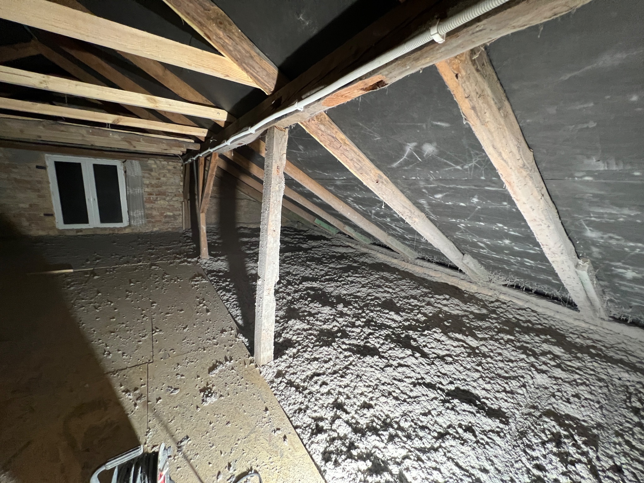 Poddasze w Białymstoku z drewnianą konstrukcją i oknem, wypełnione białą izolacją w formie luźnej wełny mineralnej, widoczne fragmenty narzędzi.