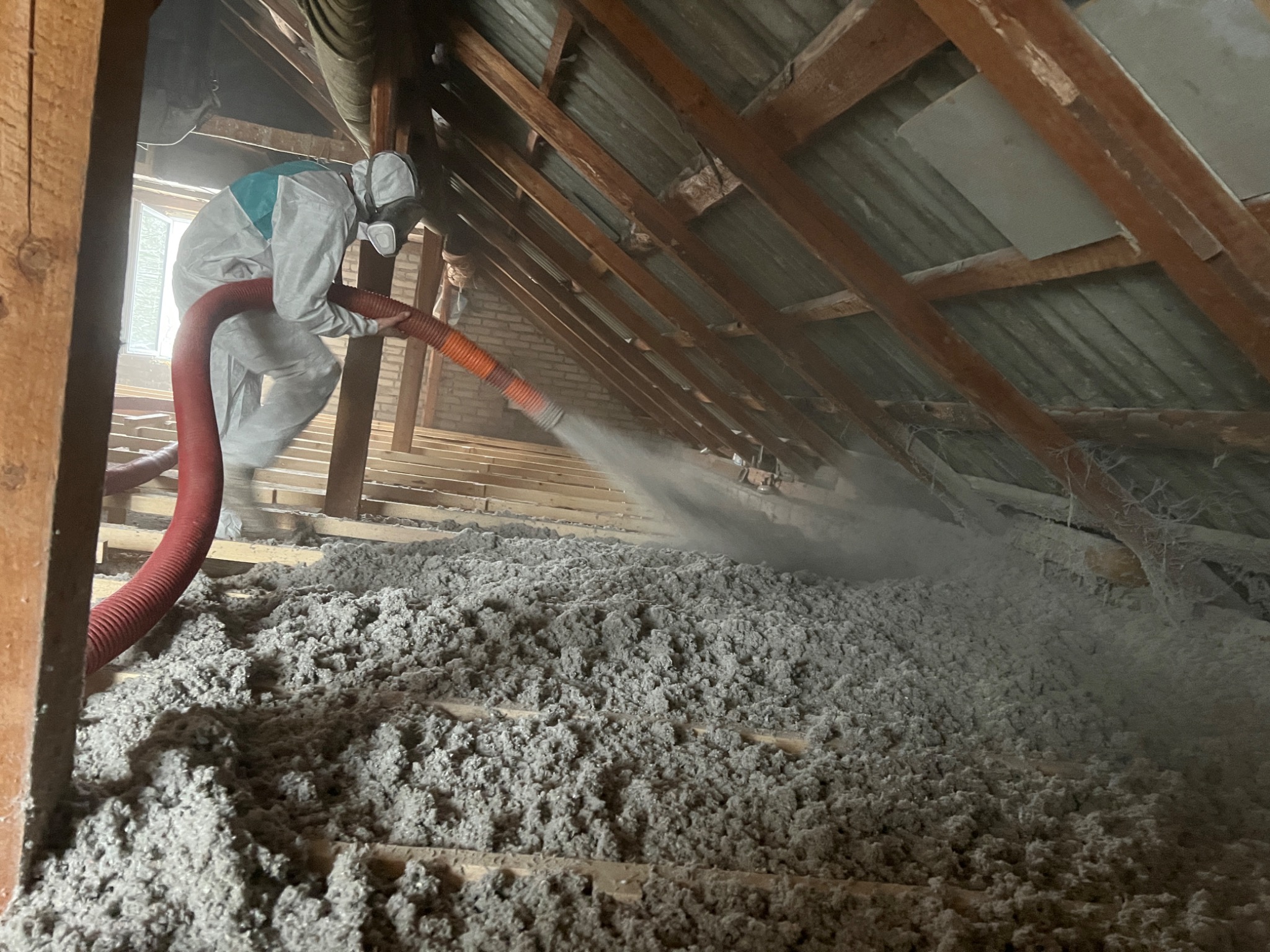 Pracownik w kombinezonie ochronnym aplikuje szarą izolację z dyszy węża na drewnianą konstrukcję poddasza, widoczne elementy więźby dachowej i fragment okna.