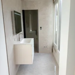 Wykończona łazienka w beżowych płytkach z wiszącą białą szafką umywalkową, lustrem i kabiną prysznicową z czarnymi ramami.