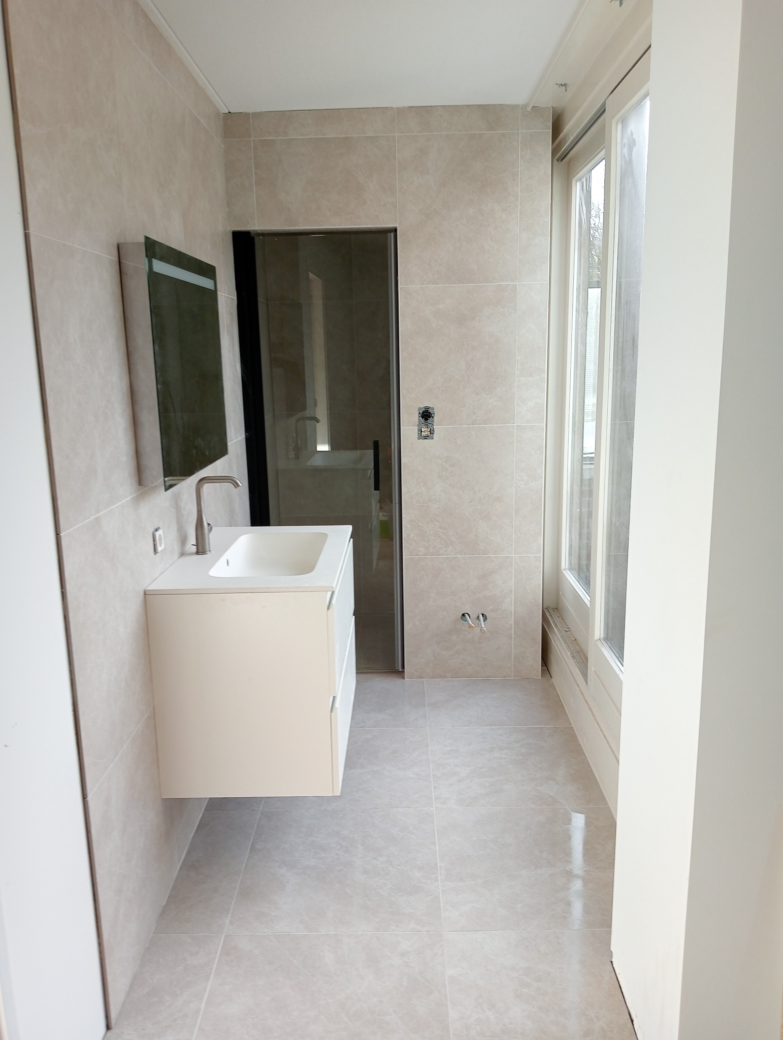 Wykończona łazienka w beżowych płytkach z wiszącą białą szafką umywalkową, lustrem i kabiną prysznicową z czarnymi ramami.