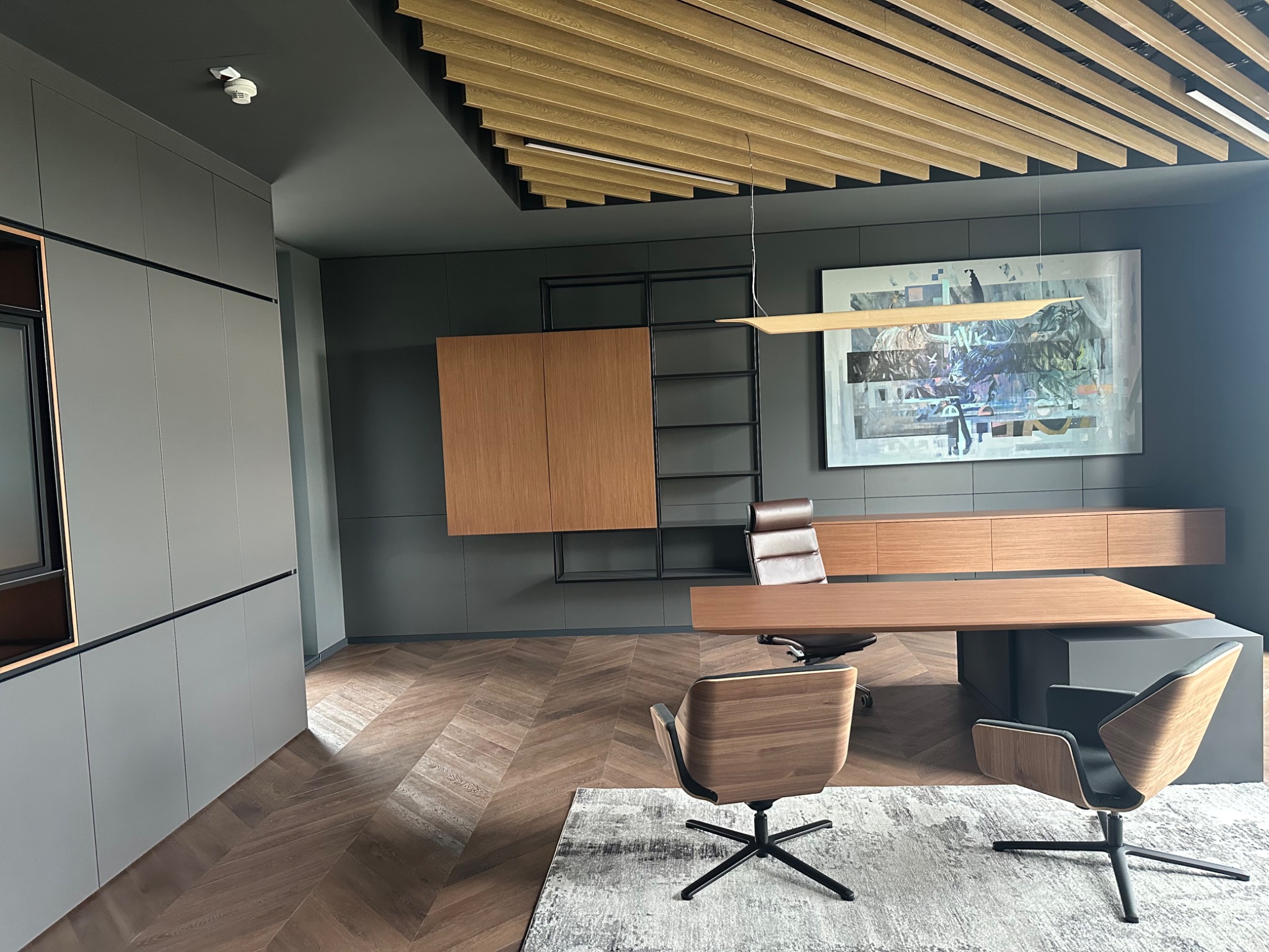 Nowoczesne biuro z drewnianą podłogą w jodełkę, szarą zabudową i designerskimi krzesłami. Na ścianie abstrakcyjny obraz, a sufit zdobi drewniana konstrukcja.