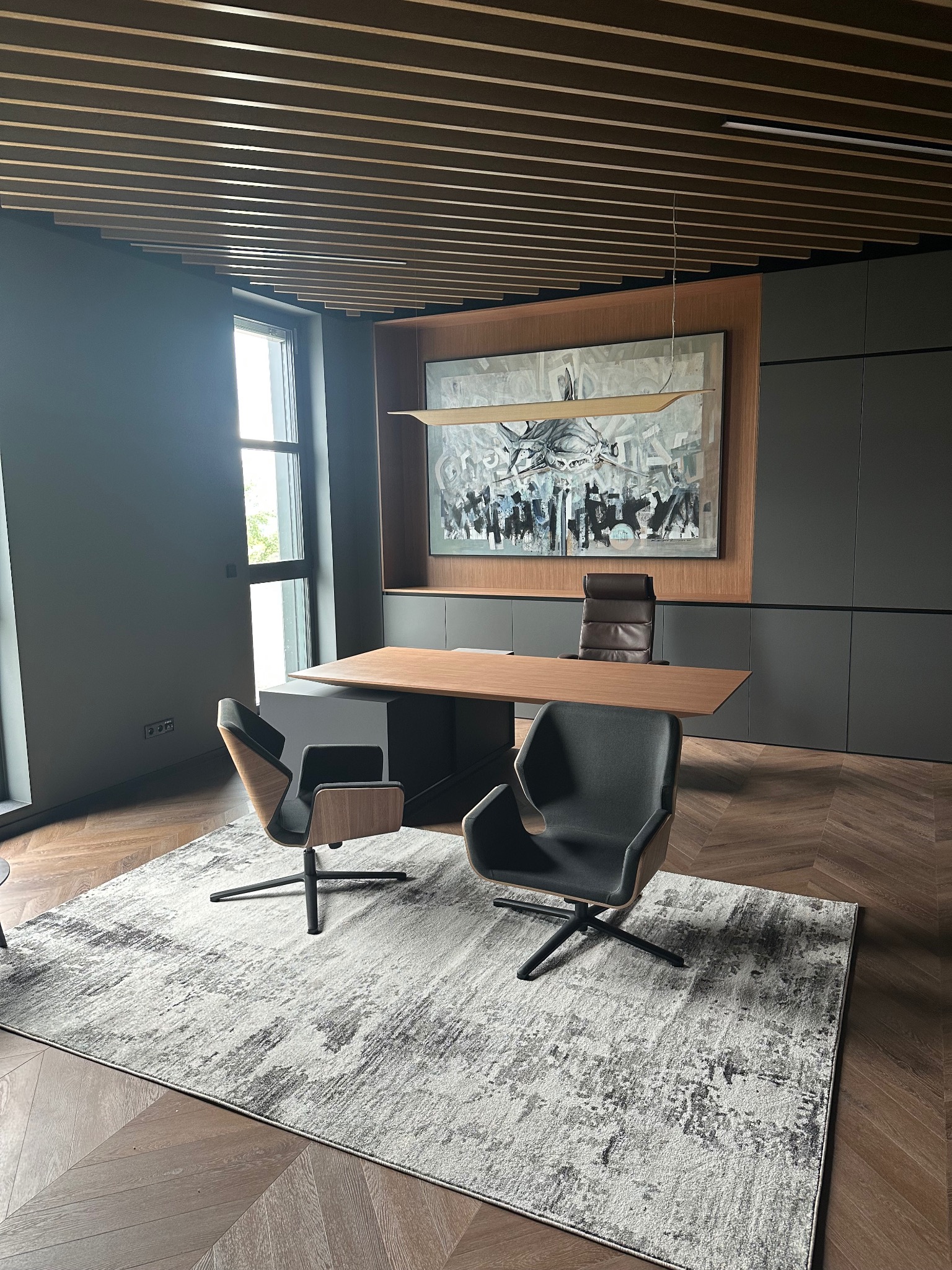 Nowoczesne biuro z drewnianym sufitem, szarą ścianą i podłogą w jodełkę. Dwa krzesła i biurko na dywanie. Obraz na ścianie i szafki w tle.
