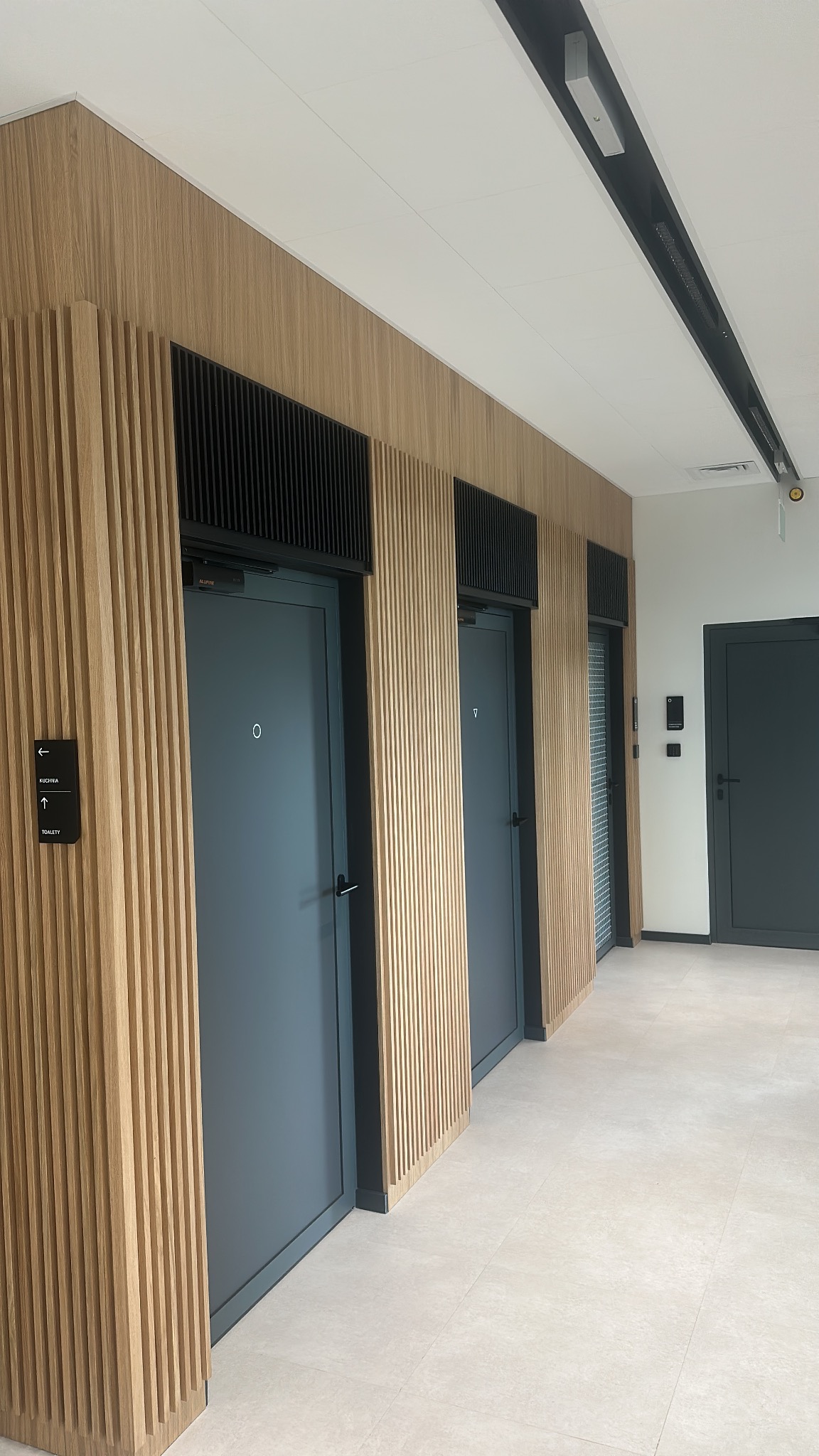 Nowoczesne wykończenie korytarza biurowego z drewnianymi panelami ściennymi i szarymi drzwiami. Widoczne detale wykończeniowe i oświetlenie sufitowe.