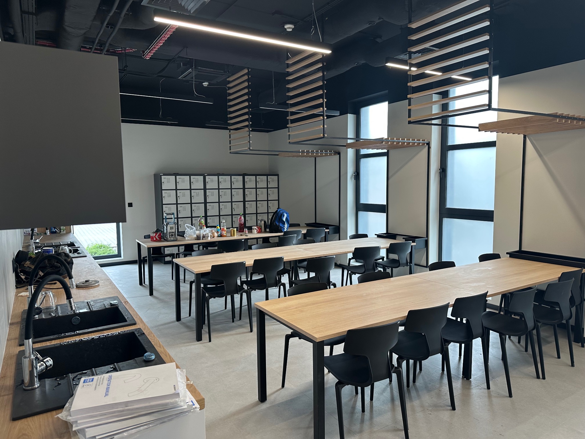 Jasne biuro po remoncie: długie stoły z krzesłami, szafki pracownicze w tle, nowoczesne oświetlenie. Widok na umywalki i okna. Minimalistyczny design.