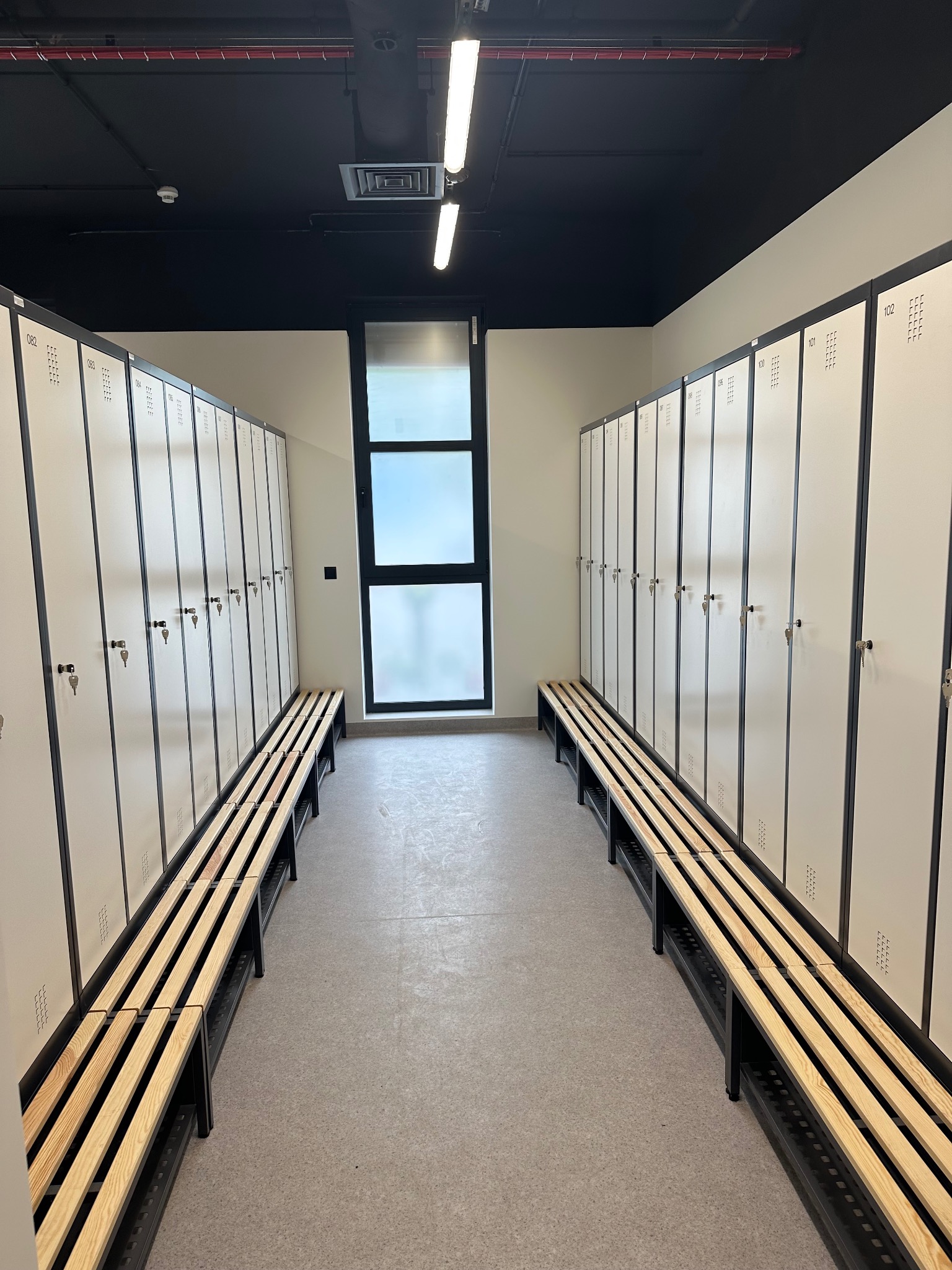 Nowoczesna szatnia z rzędami białych szafek z numerami i drewnianymi ławkami. Minimalistyczny design, czarne sufity, jasne oświetlenie i okno w tle.