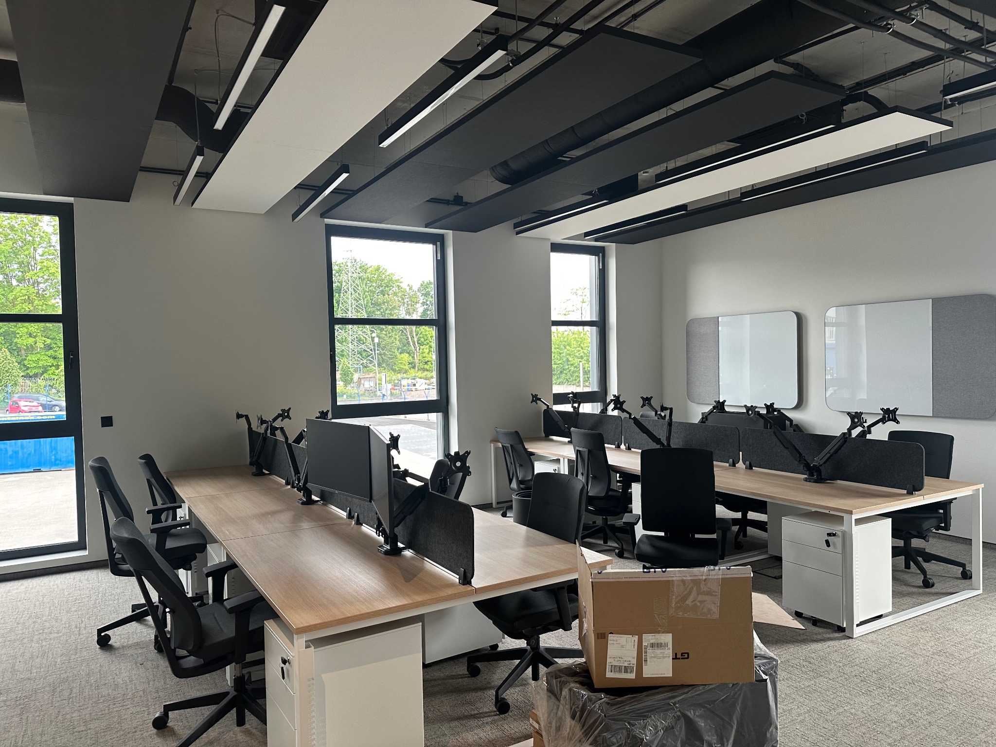 Jasne biuro po remoncie: rzędy biurek z krzesłami, duże okna, nowoczesne oświetlenie sufitowe. Kartonowe pudła w rogu sugerują świeże wykończenie.