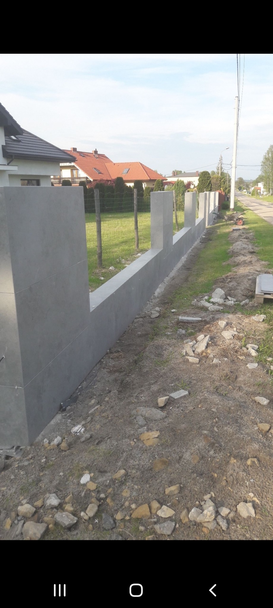 Nowoczesny, szary, betonowy płot panelowy w trakcie budowy, z widocznymi elementami konstrukcyjnymi i otoczeniem posesji. Ujęcie z boku, w dziennym świetle.