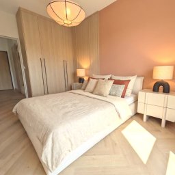 Sopata Studio Design Katarzyna Sopata - Elegancka sypialnia z beżowym łóżkiem, szafą z jasnego drewna, ścianą w odcieniu terakoty i podłogą w jodełkę. Unikalna lampa sufitowa, promienie słońca na parkiecie.