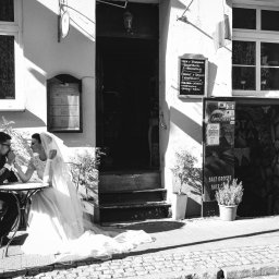 Karina Derda - Czarno-białe zdjęcie pary młodej siedzącej przy stoliku na zewnątrz restauracji w słoneczny dzień, panna młoda w welonie i sukni, pan młody w garniturze, trzymający ją za rękę, na tle ulicy...