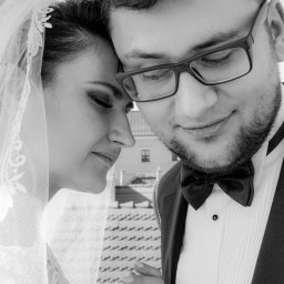 Karina Derda - Czarno-białe zbliżenie młodej pary: panna młoda w welonie opiera głowę o głowę pana młodego w muszce i okularach, na tle dachu z dachówkami.