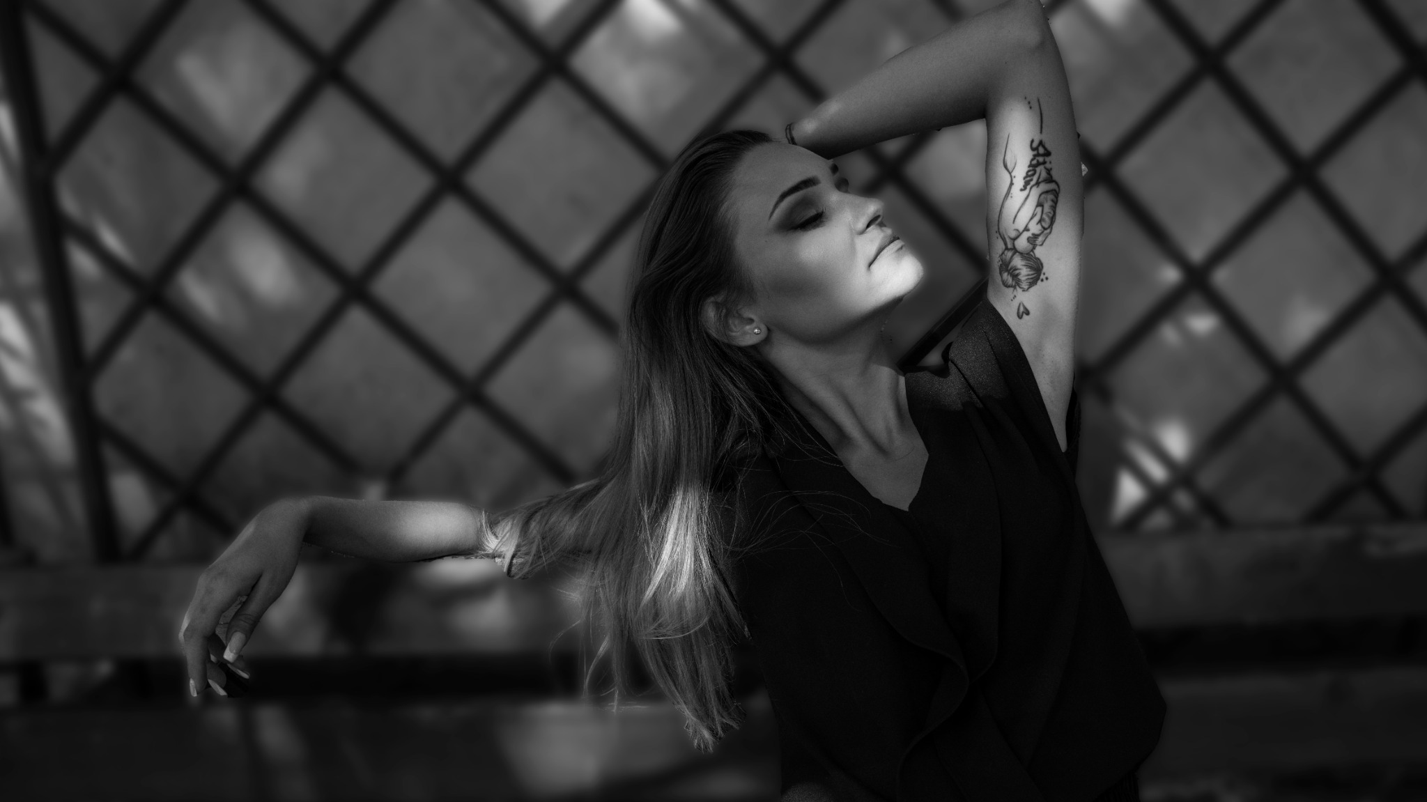 Czarno-białe zdjęcie kobiety z tatuażem na ramieniu, z zamkniętymi oczami, wystawiona na promienie słoneczne, na tle kratownicy.