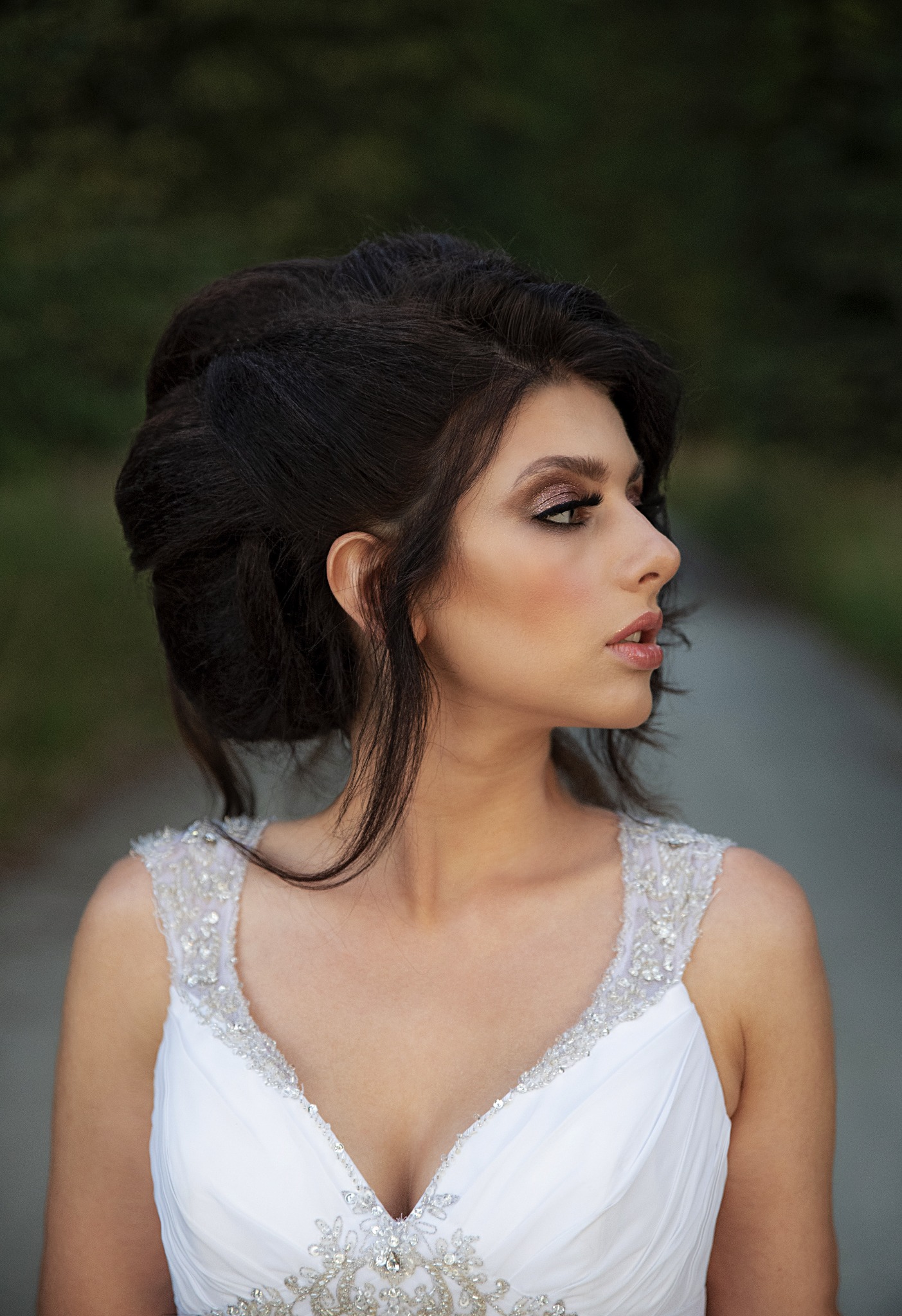 Portret panny młodej w białej sukni z kryształkami, z upiętymi ciemnymi włosami i wyrazistym makijażem, profil na tle rozmytej zieleni.