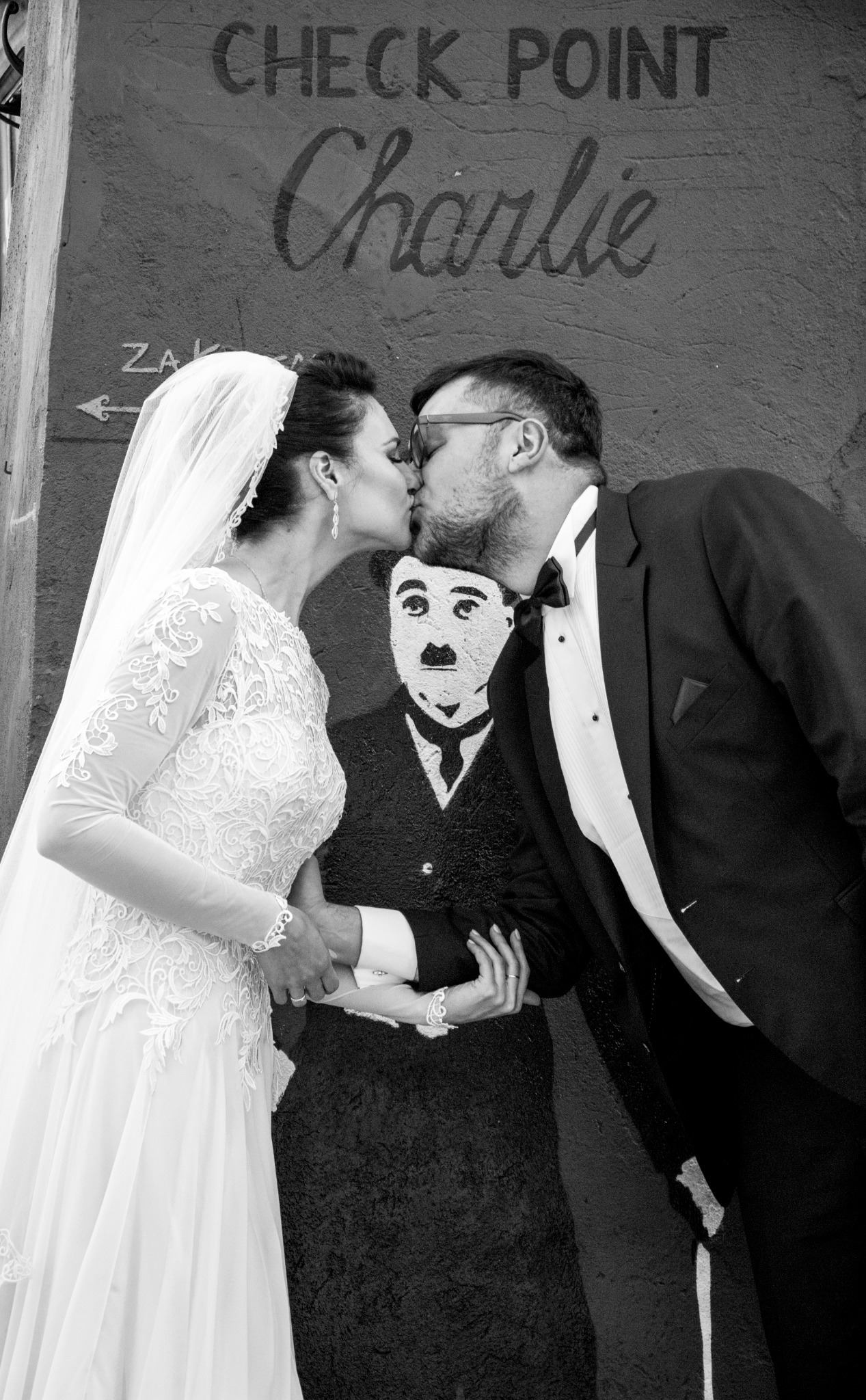 Czarno-białe zdjęcie całującej się pary młodej na tle ściany z napisem 'Check Point Charlie' i wizerunkiem Charliego Chaplina.