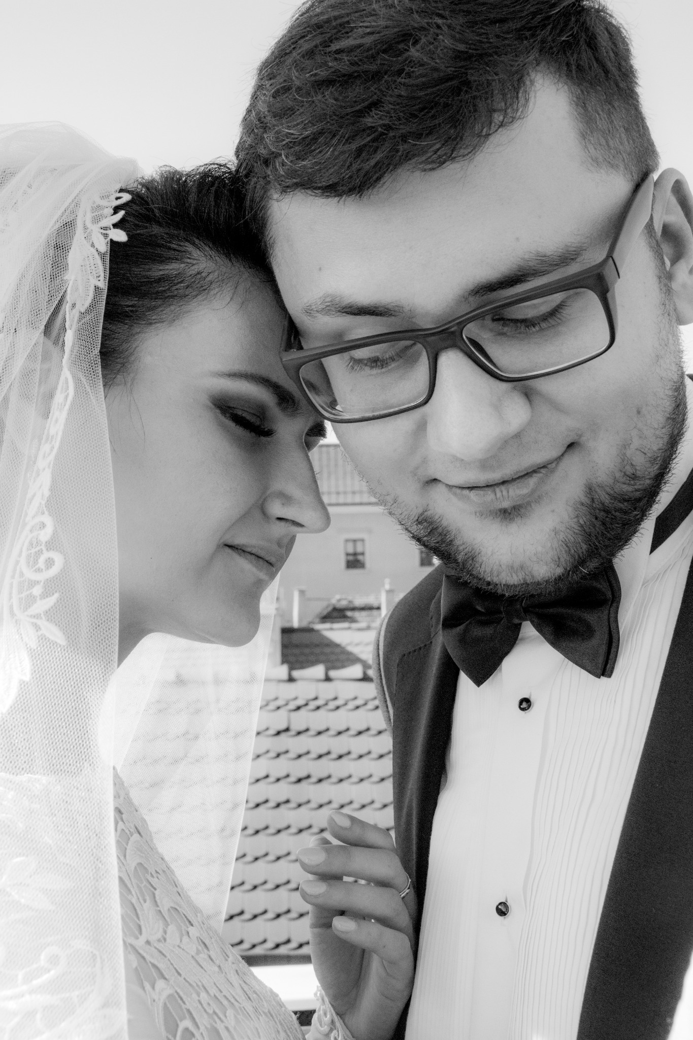 Czarno-białe zbliżenie młodej pary: panna młoda w welonie opiera głowę o głowę pana młodego w muszce i okularach, na tle dachu z dachówkami.