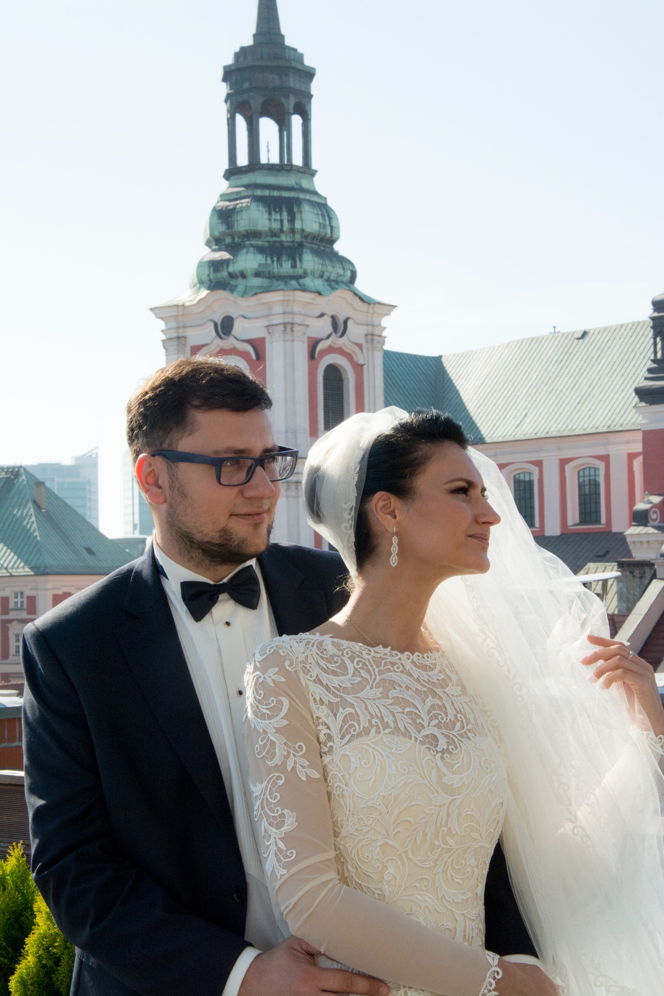 Para młoda w eleganckich strojach, Panna Młoda w koronkowej sukni z długim rękawem i welonem, Pan Młody w smokingu z muszką, w tle zabytkowy budynek z wieżą