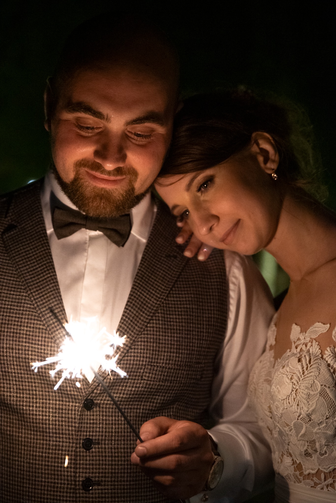 Uśmiechnięta para w ciemności, mężczyzna w kraciastej kamizelce trzyma zapaloną zimną iskrę, kobieta opiera głowę na jego ramieniu, widoczny fragment koronkowej sukni.