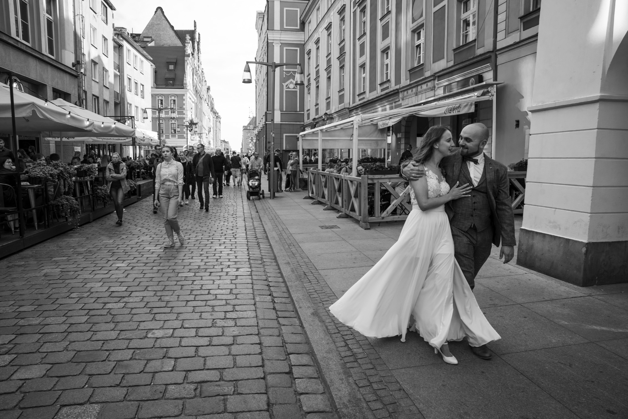 Czarno-białe zdjęcie pary młodej spacerującej po brukowanej ulicy w europejskim mieście, panna młoda w koronkowej sukni z rozkloszowanym dołem, pan młody w tweedowym garniturze z muszką, w tle...