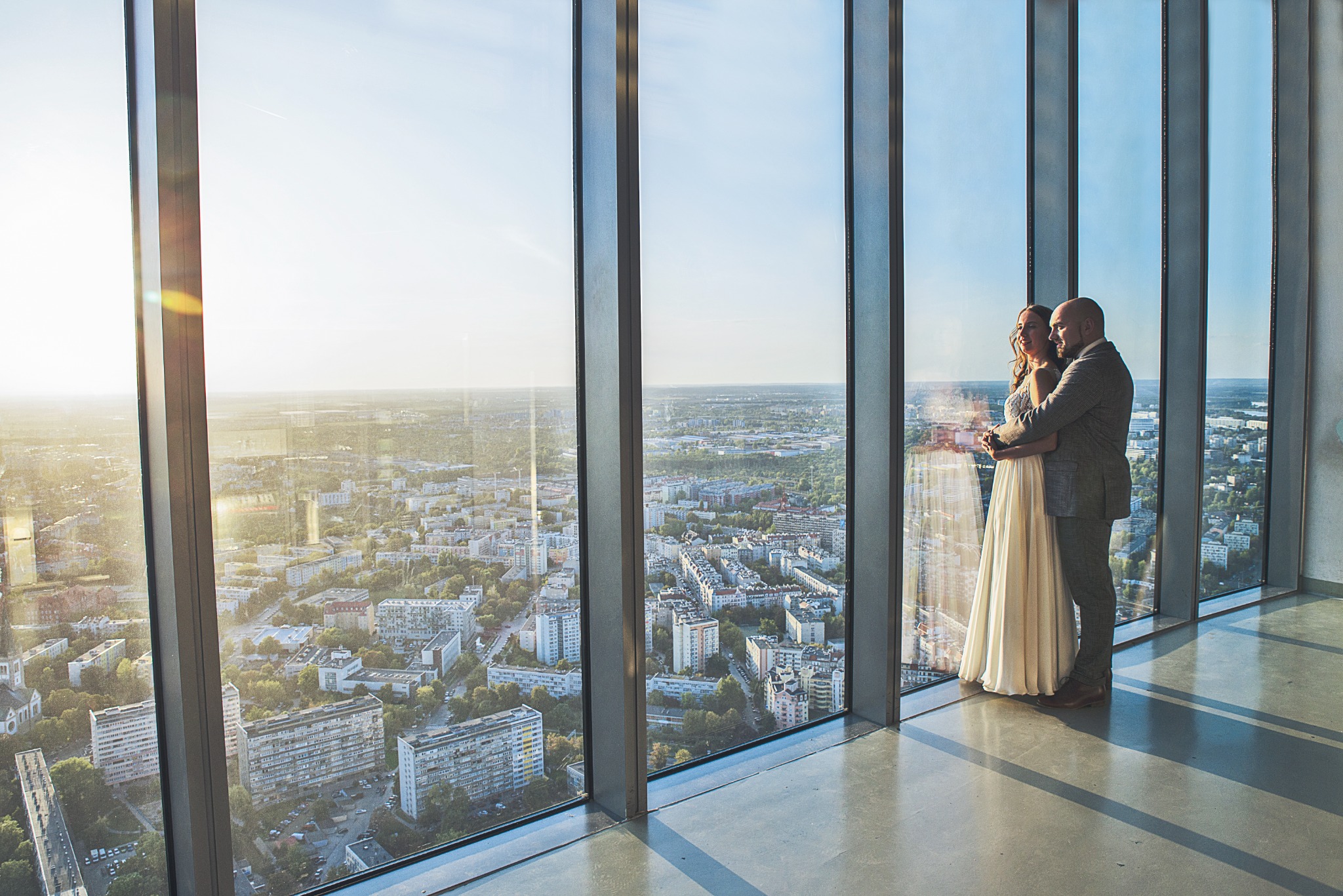Para młoda w objęciach na tle panoramicznego okna z widokiem na miasto o zachodzie słońca, ona w sukni ślubnej z koronkową górą, on w szarym garniturze w kratę.