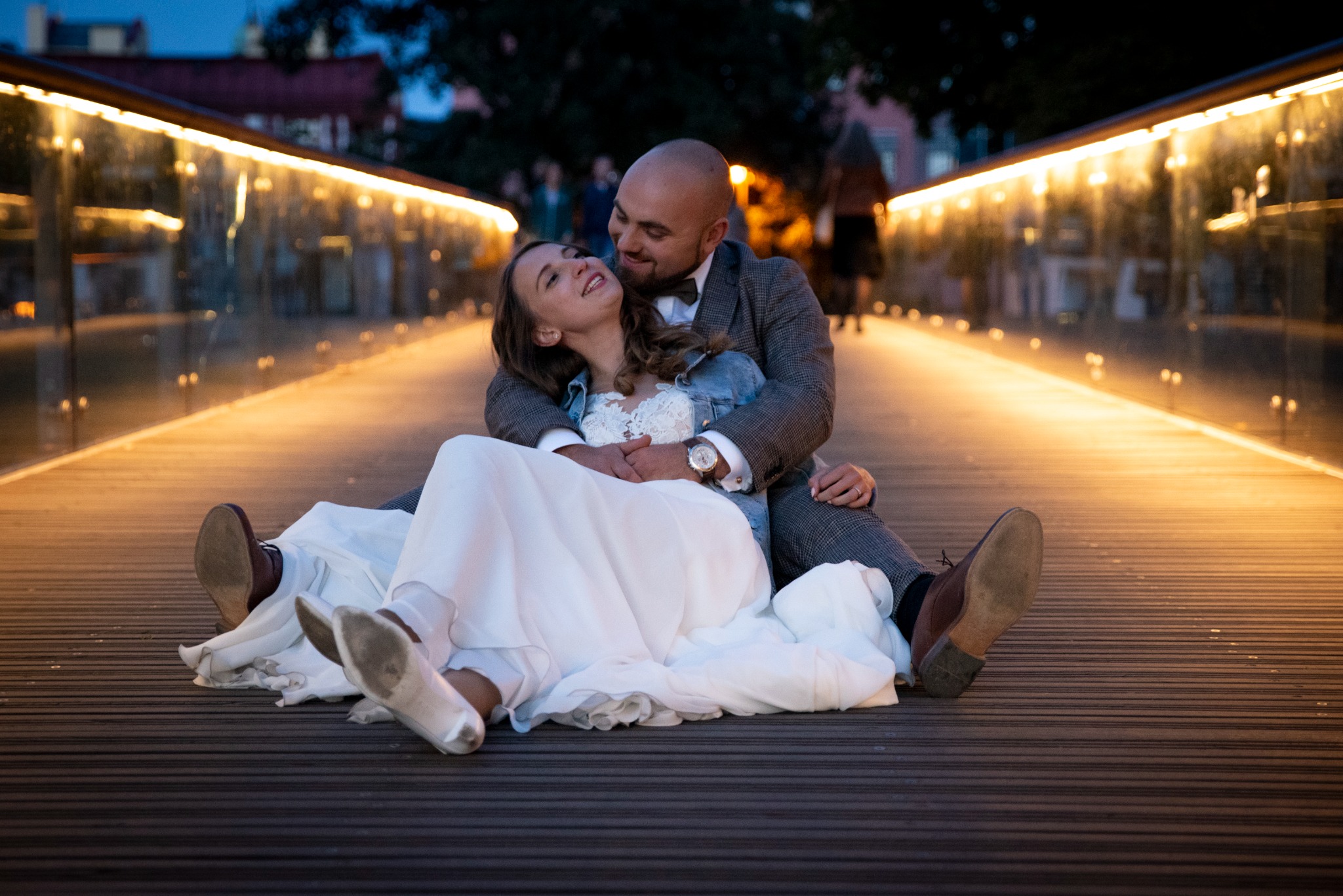 Para młoda siedzi na oświetlonym moście, panna młoda w zwiewnej sukni i jeansowej kurtce, pan młody w tweedowej marynarce i muszce, oboje uśmiechnięci.