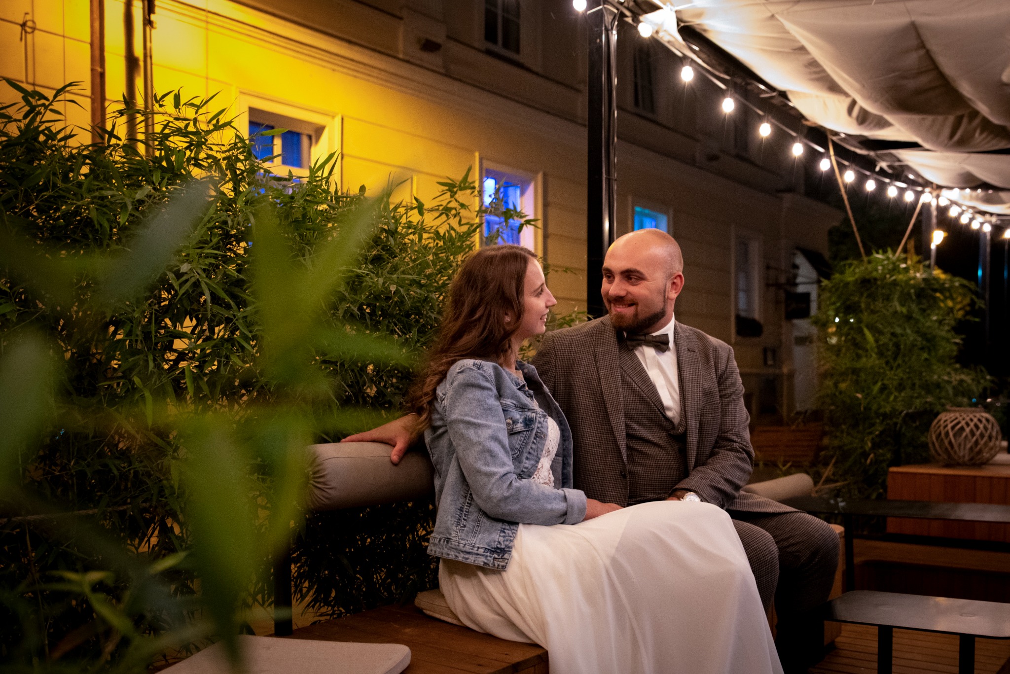 Uśmiechnięta para w strojach ślubnych siedzi na ławce w ogrodowej scenerii pod girlandami światełek, panna młoda w jeansowej kurtce.