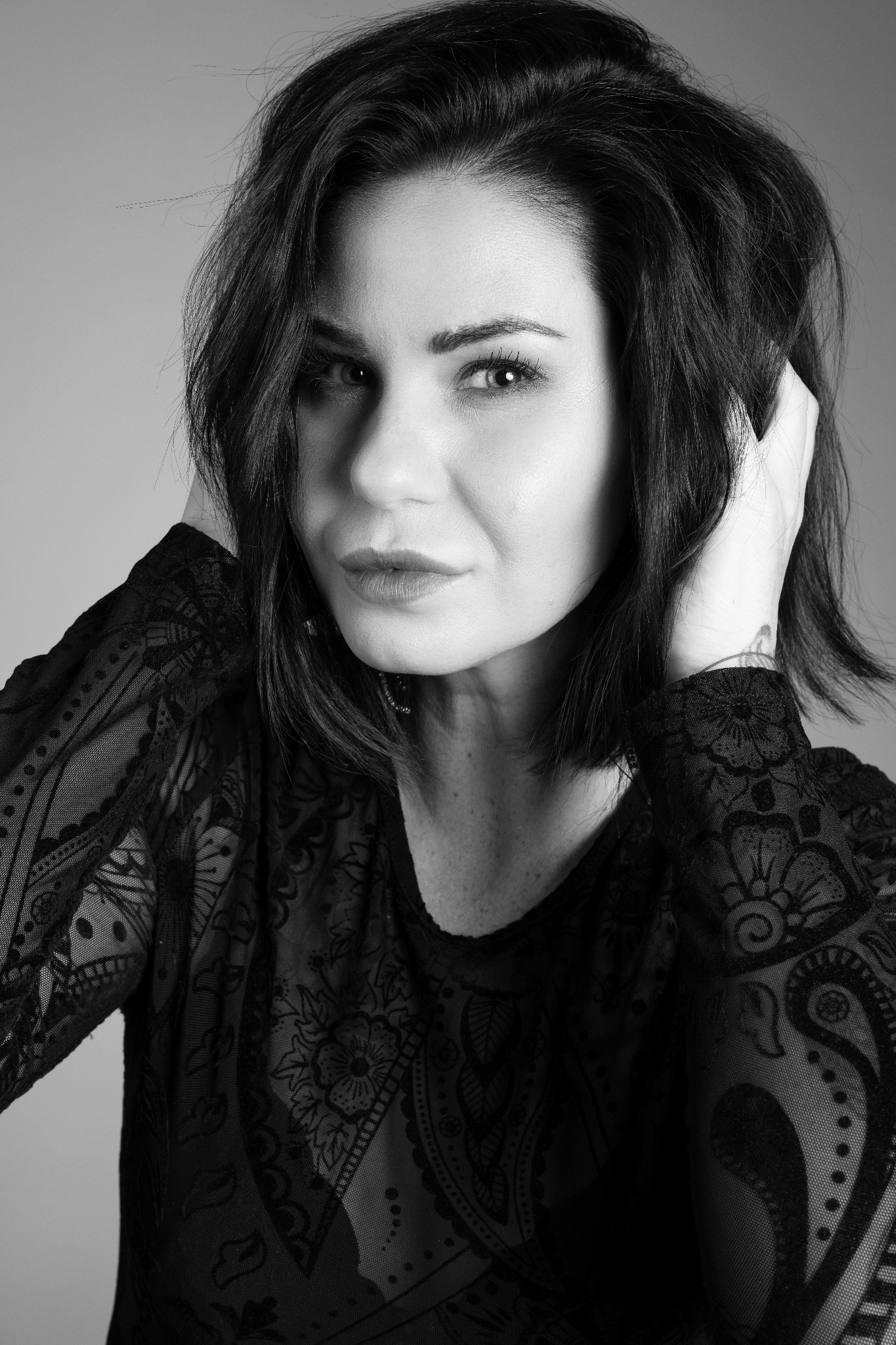 Portret kobiety w czarno-białej tonacji, ubrana w koronkową bluzkę, z rękami uniesionymi do włosów, patrząca prosto w obiektyw z subtelnym uśmiechem.