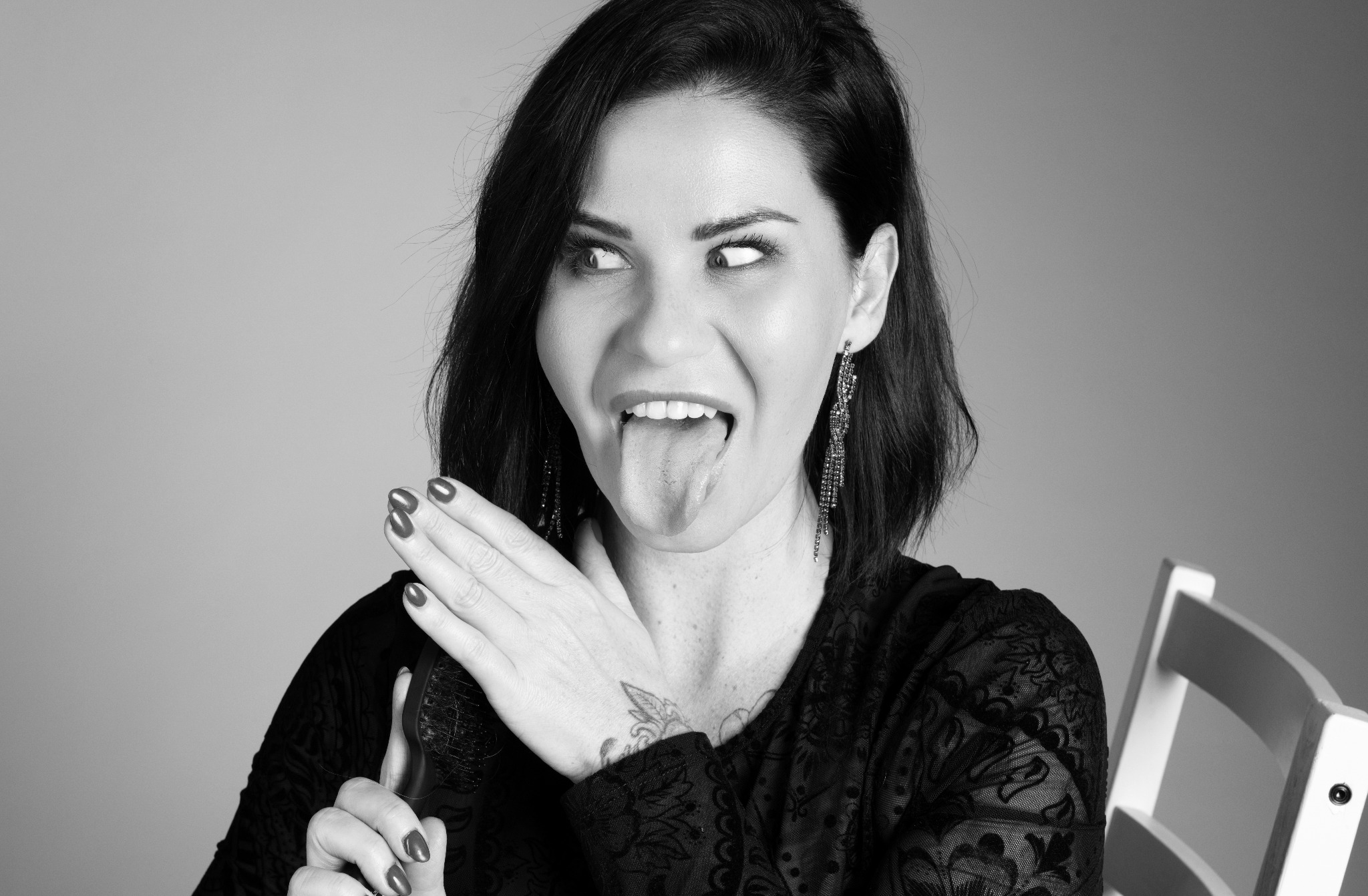 Czarno-białe zdjęcie portretowe kobiety z rozpuszczonymi, ciemnymi włosami, wystawiającej język i zezującej, ubrana w koronkową bluzkę i długie kolczyki, z tatuażem na dekolcie.